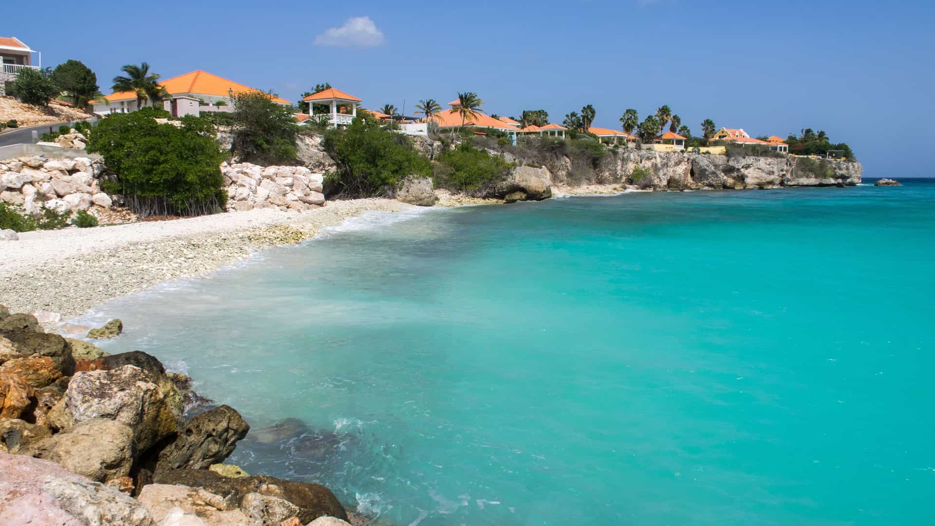 A scenic coastal view in Curaçao featuring houses with orange roofs on a cliffside overlooking a rocky beach with beautiful turquoise Caribbean water.