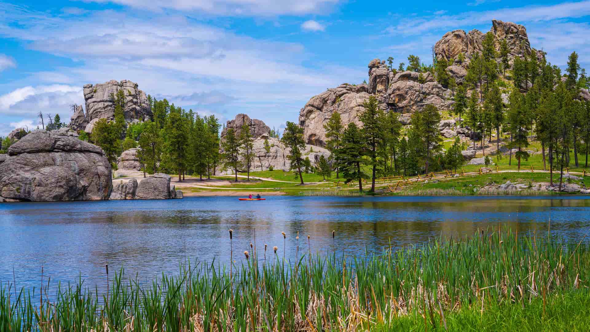 "A tranquil lake with lush reeds and grasses in the foreground, reflecting the surrounding granite rock formations and ponderosa pine trees, with two people in a red kayak on the water in Custer State Park, South Dakota. "