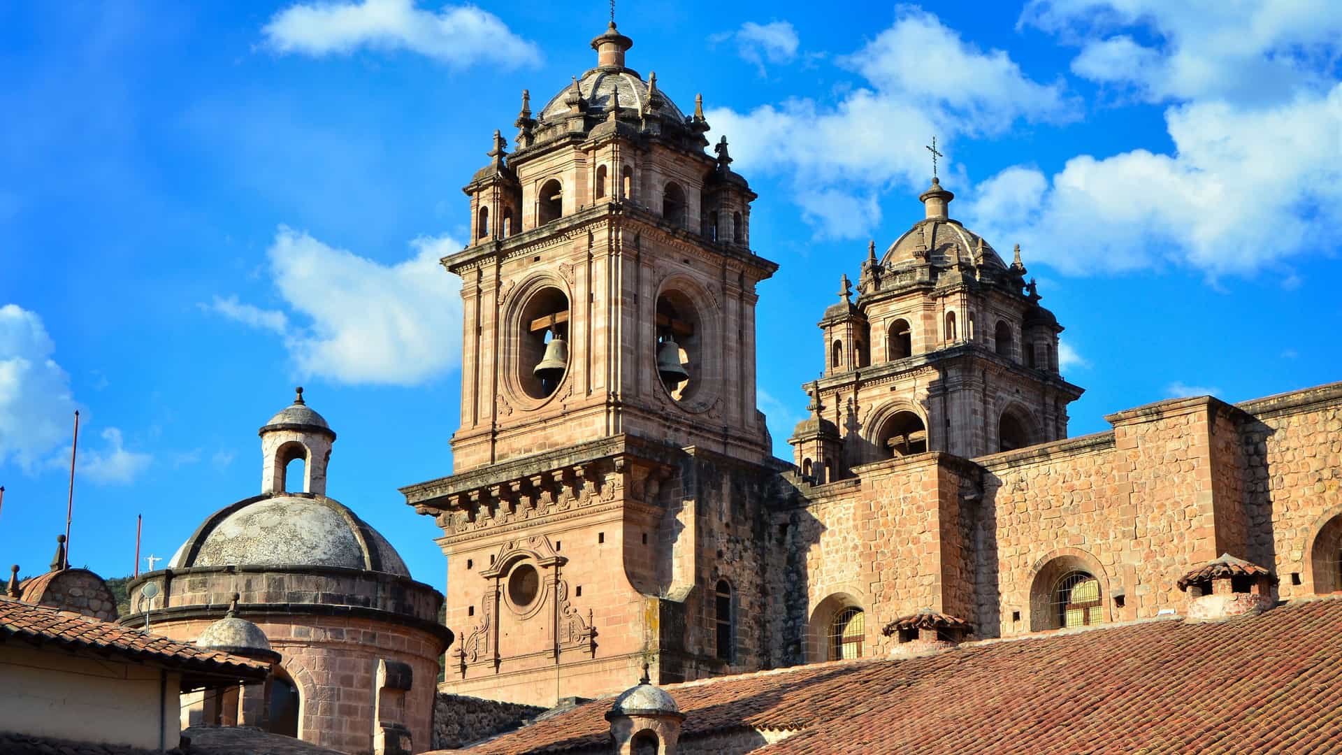 The ornate Baroque bell towers and domes of a historic stone cathedral in Cuzco, Peru, set against a bright blue sky with clouds.