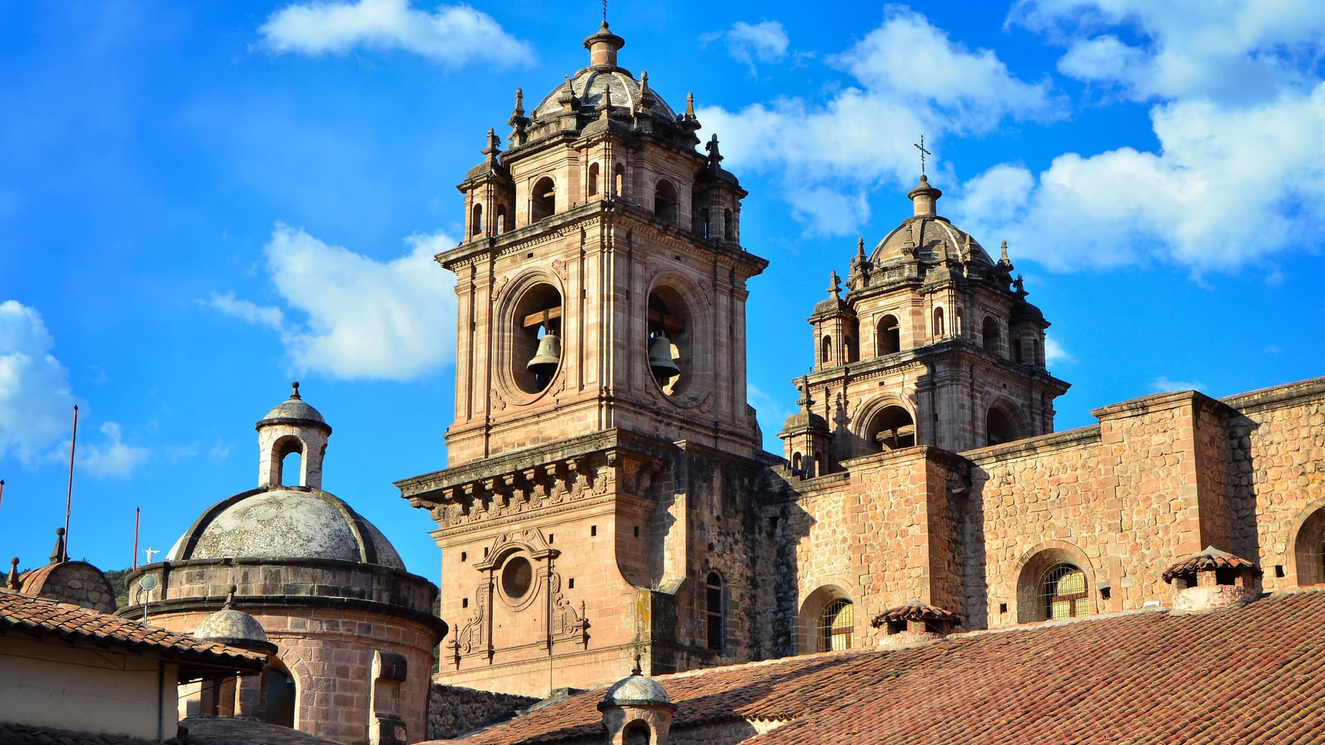 The ornate Baroque bell towers and domes of a historic stone cathedral in Cuzco, Peru, set against a bright blue sky with clouds.