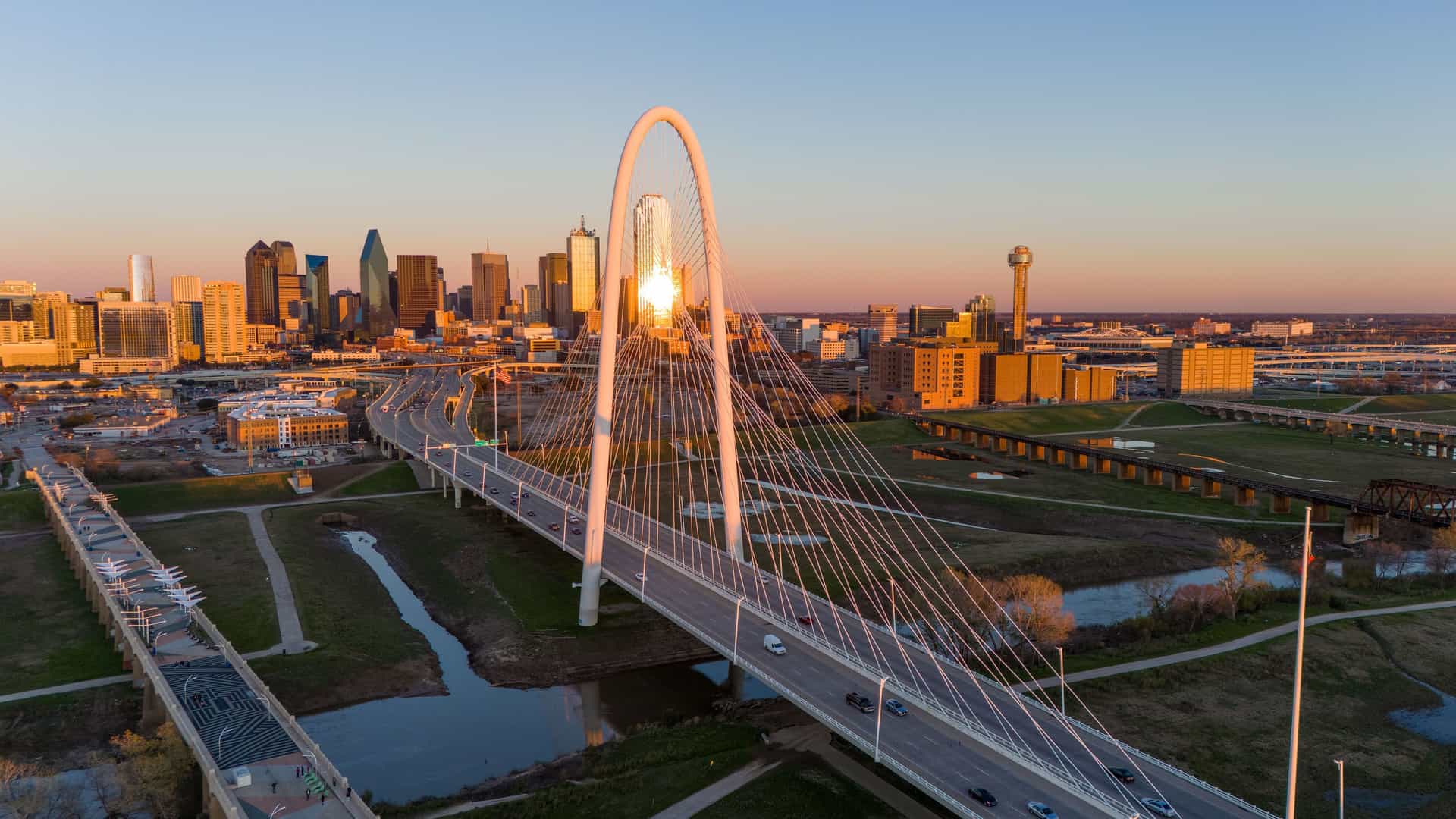 "An aerial shot of the Margaret Hunt Hill Bridge spanning the Trinity River, with cars driving across it at sunset, and the modern skyline of downtown Dallas, Texas, in the background. "