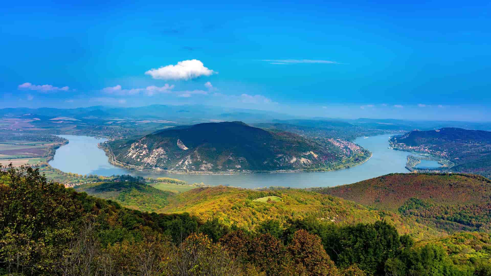 A wide-angle panoramic view of the dramatic horseshoe-shaped Danube Bend, a famous landmark in Hungary, surrounded by autumn-colored forests and a charming town.
