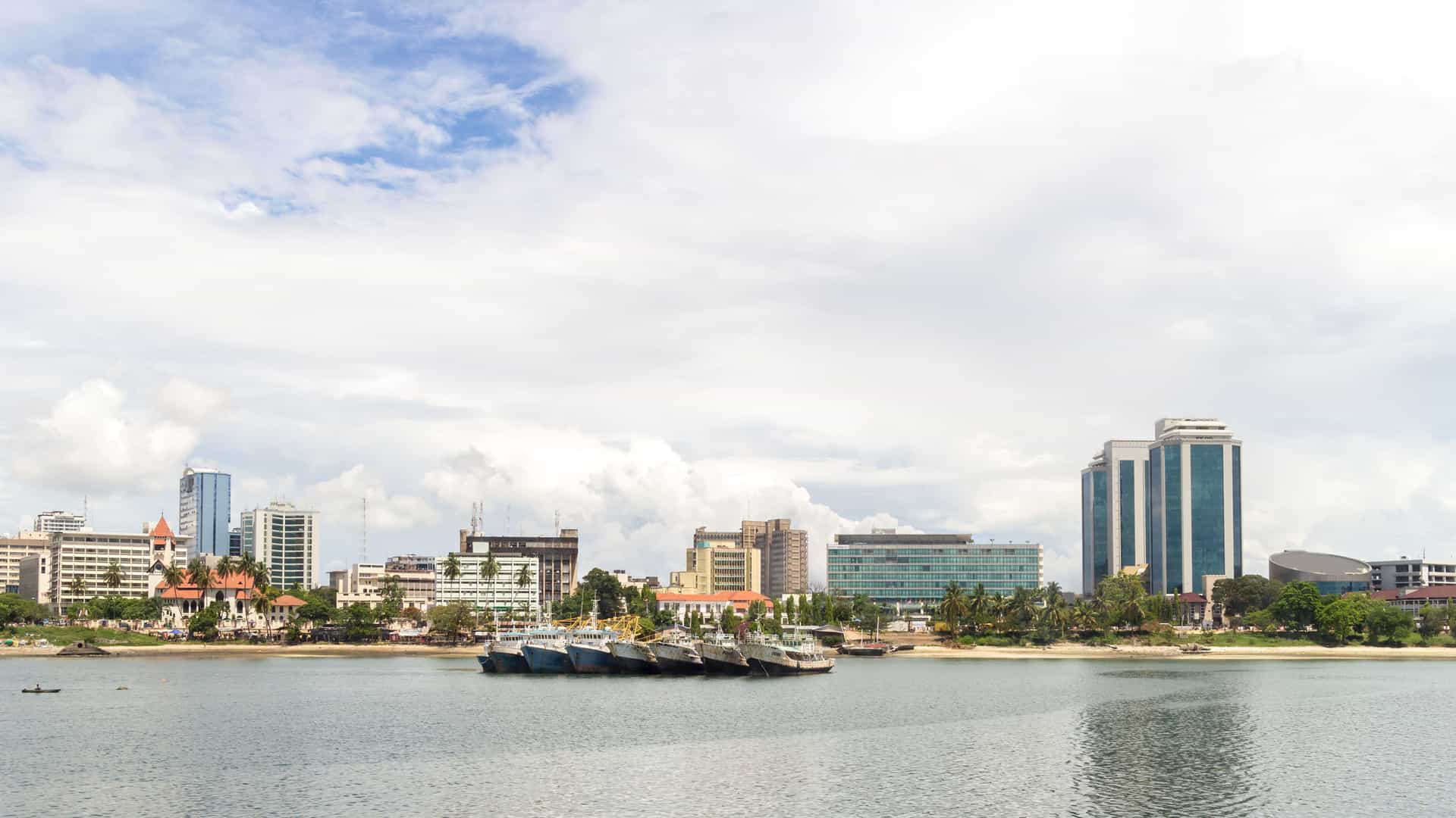 A view of the Dar es Salaam skyline in Tanzania from the water, showing modern office buildings behind traditional wooden boats moored in the harbor.