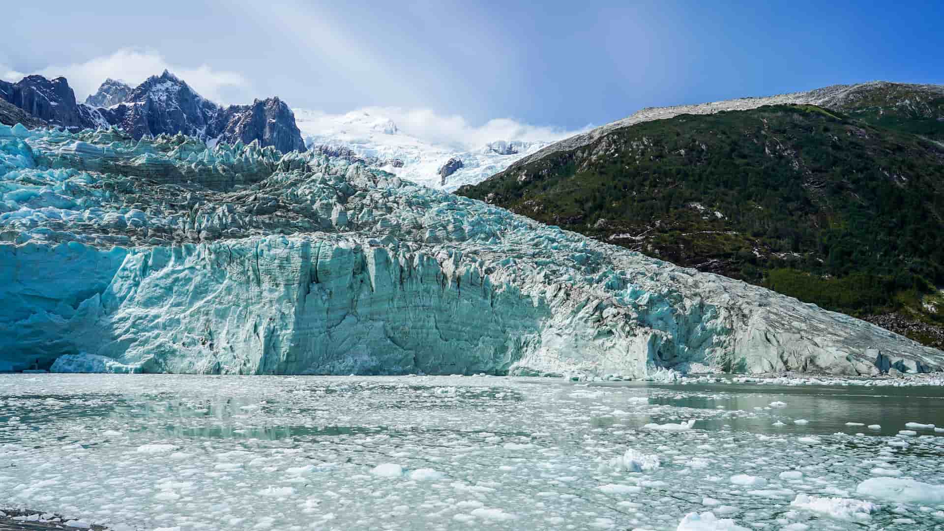 A close-up view of the massive blue-toned D'Agostini Glacier in the Darwin Channel, with a backdrop of snow-capped mountains and green slopes.