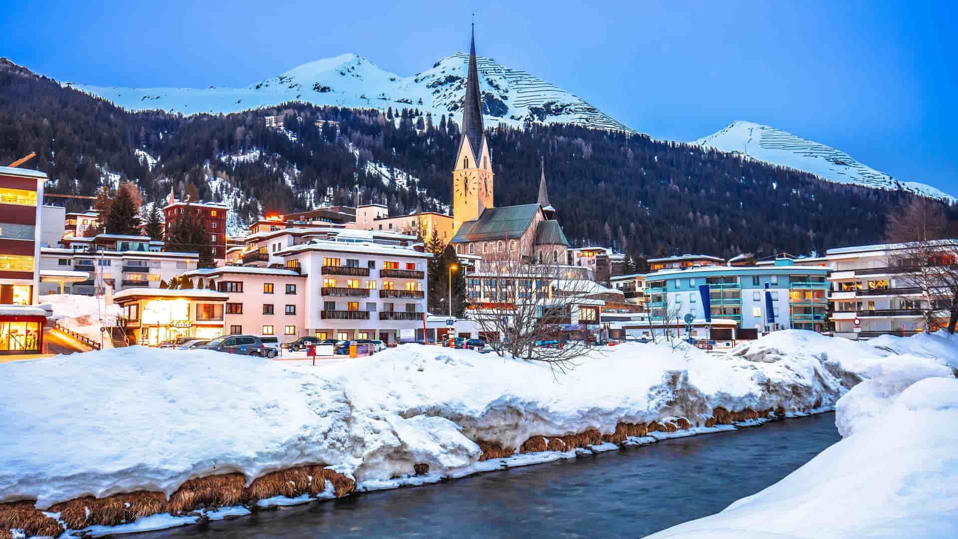 Snow-covered village of Davos, Switzerland, with a flowing river and St. Johann Church spire against the backdrop of the Swiss Alps.