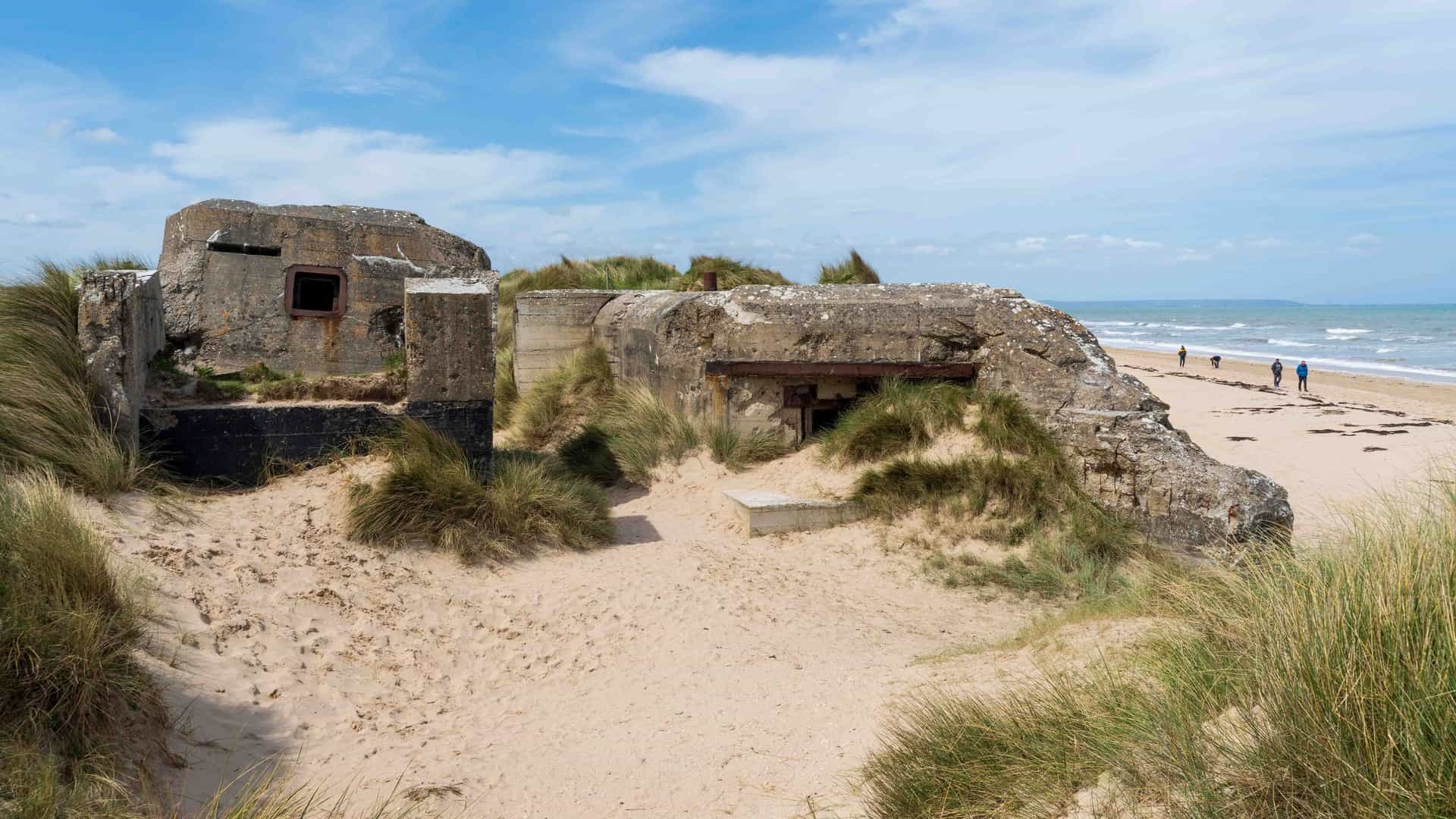A wide-angle view of a weathered German bunker on Omaha Beach in Normandy, France, a significant D-Day landing site, with beachgoers in the distance.
