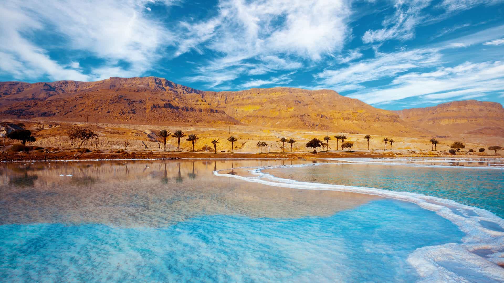 A panoramic view of the Dead Sea in Jordan, with bright blue, reflective water and unique salt formations set against arid, rugged mountains.