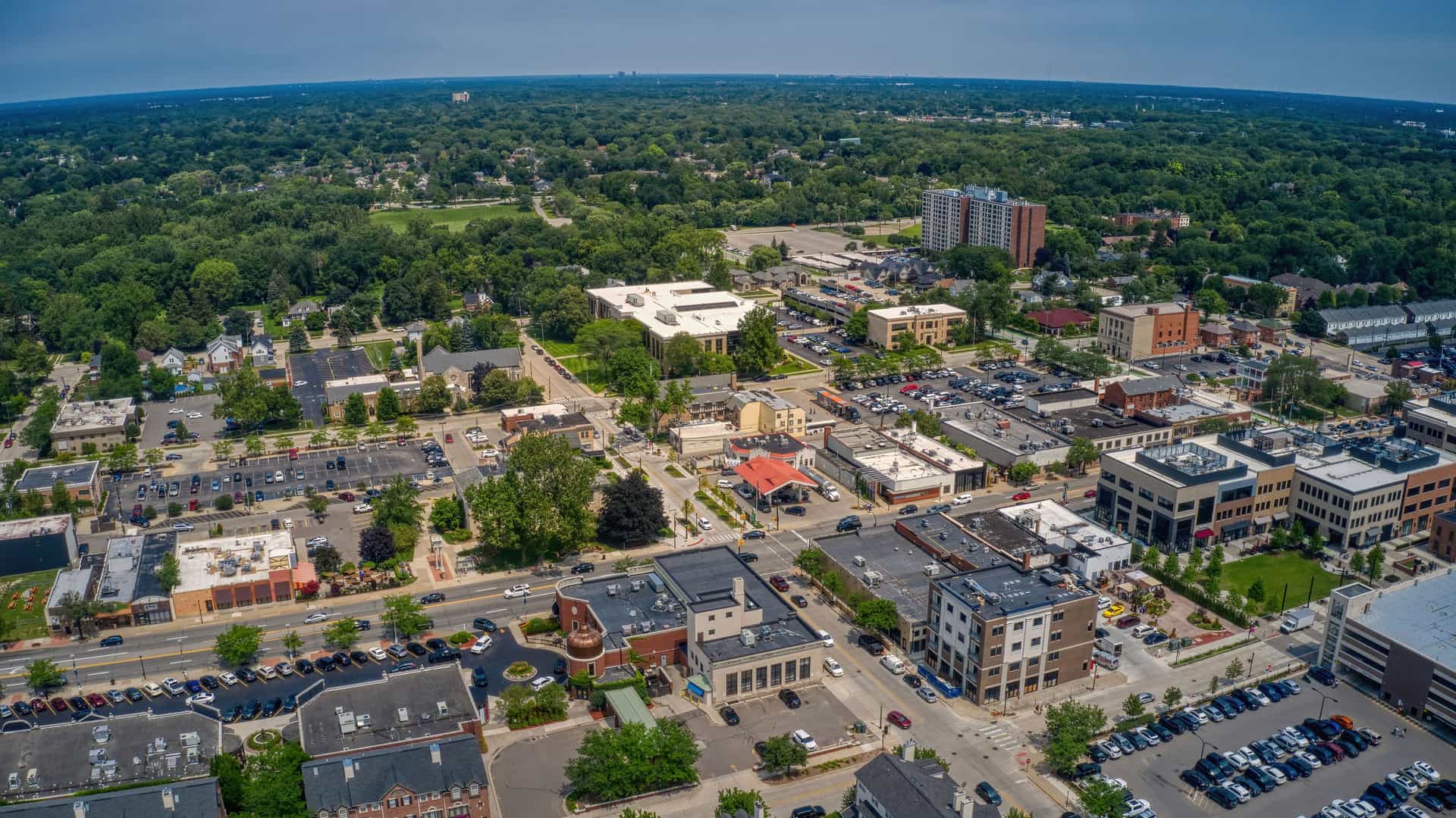 "An aerial view of downtown Dearborn, Michigan, showcasing a mix of modern and historic buildings, commercial areas, and residential neighborhoods surrounded by the dense green trees of the city. "