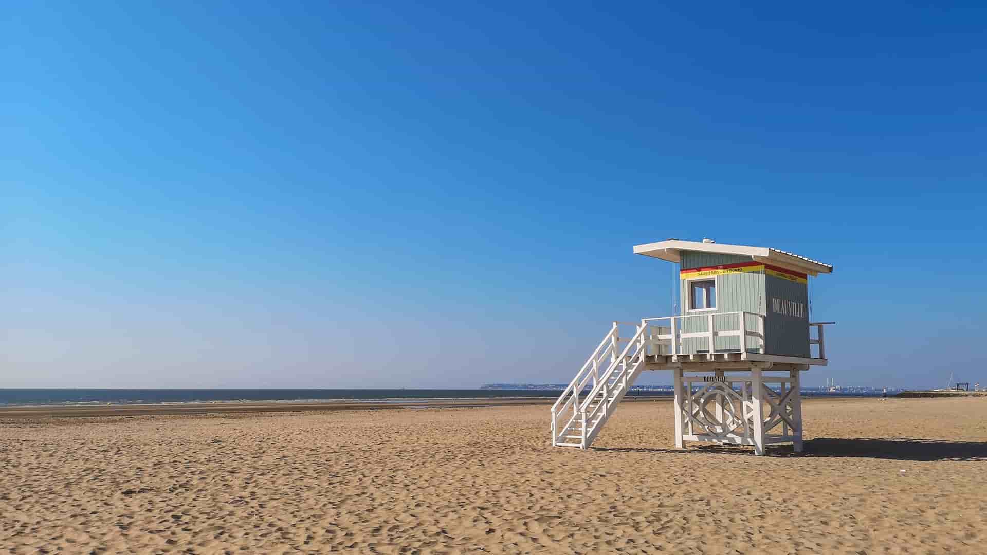 "A small, light green and white lifeguard hut with a wooden staircase sits on the wide, sandy beach of Deauville, France, with a calm blue sea and a clear sky stretching to the horizon. "