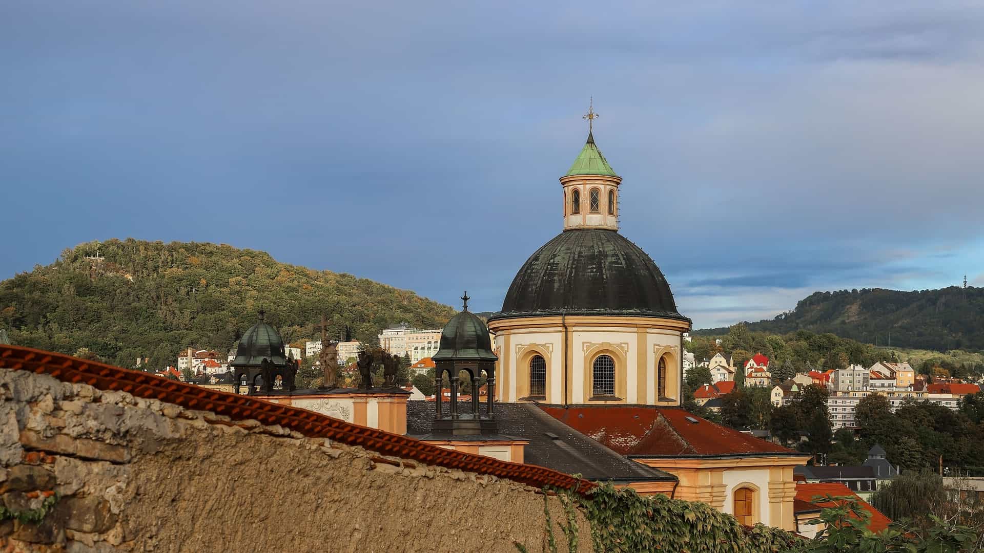 An exterior view of the Baroque-style domed Church of the Holy Cross at Děčín Castle, overlooking the historic town and forested hills.