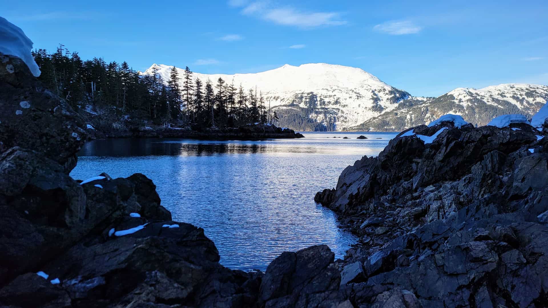 A scenic view of the calm waters of Decision Passage, with a snow-dusted, forested shoreline and majestic snow-capped mountains in the background.