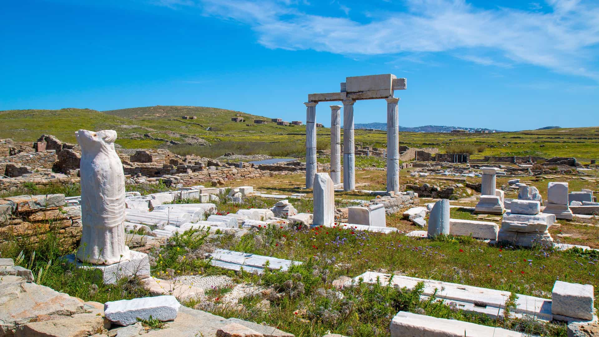 "A scenic shot of the ancient archaeological ruins on the Greek island of Delos, with broken marble columns and statues scattered across a grassy field under a bright blue sky. "