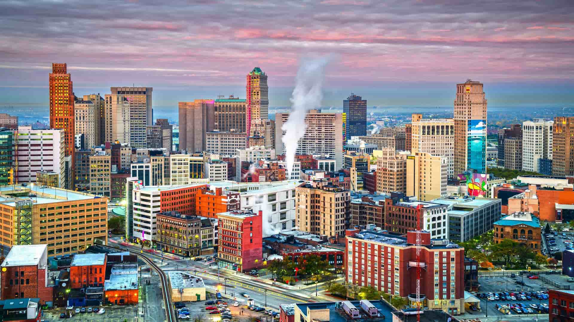An aerial view of the Detroit skyline at dusk, with its modern skyscrapers and historic buildings glowing under a vibrant pink and purple sky.