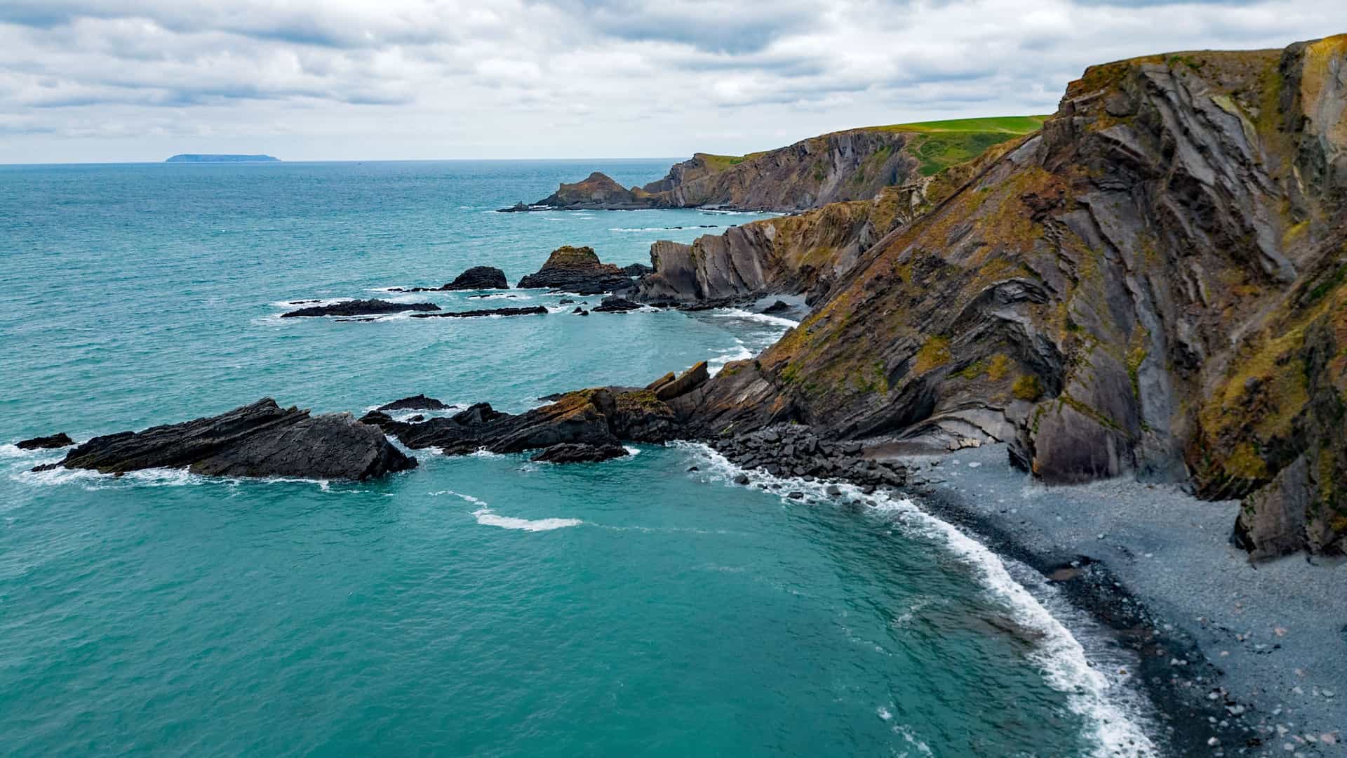 An aerial view of the dramatic and rugged coastline of Devon, England, showcasing its unique, folded rock formations, steep cliffs, and the turquoise waters of the Atlantic Ocean.