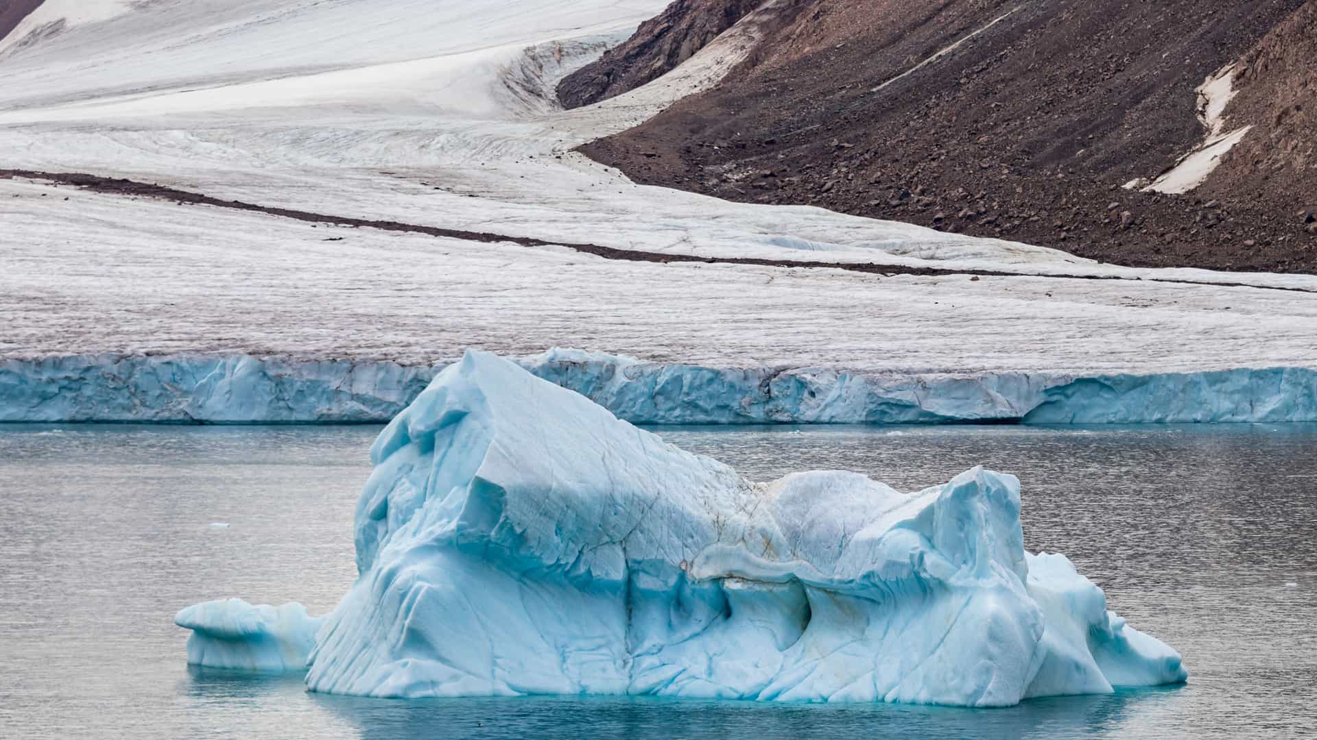 "A large, blue iceberg floating in the frigid waters of the Arctic Ocean, with a massive glacier and a dark brown mountain in the background on Diana Island, Nunavut, Canada. "