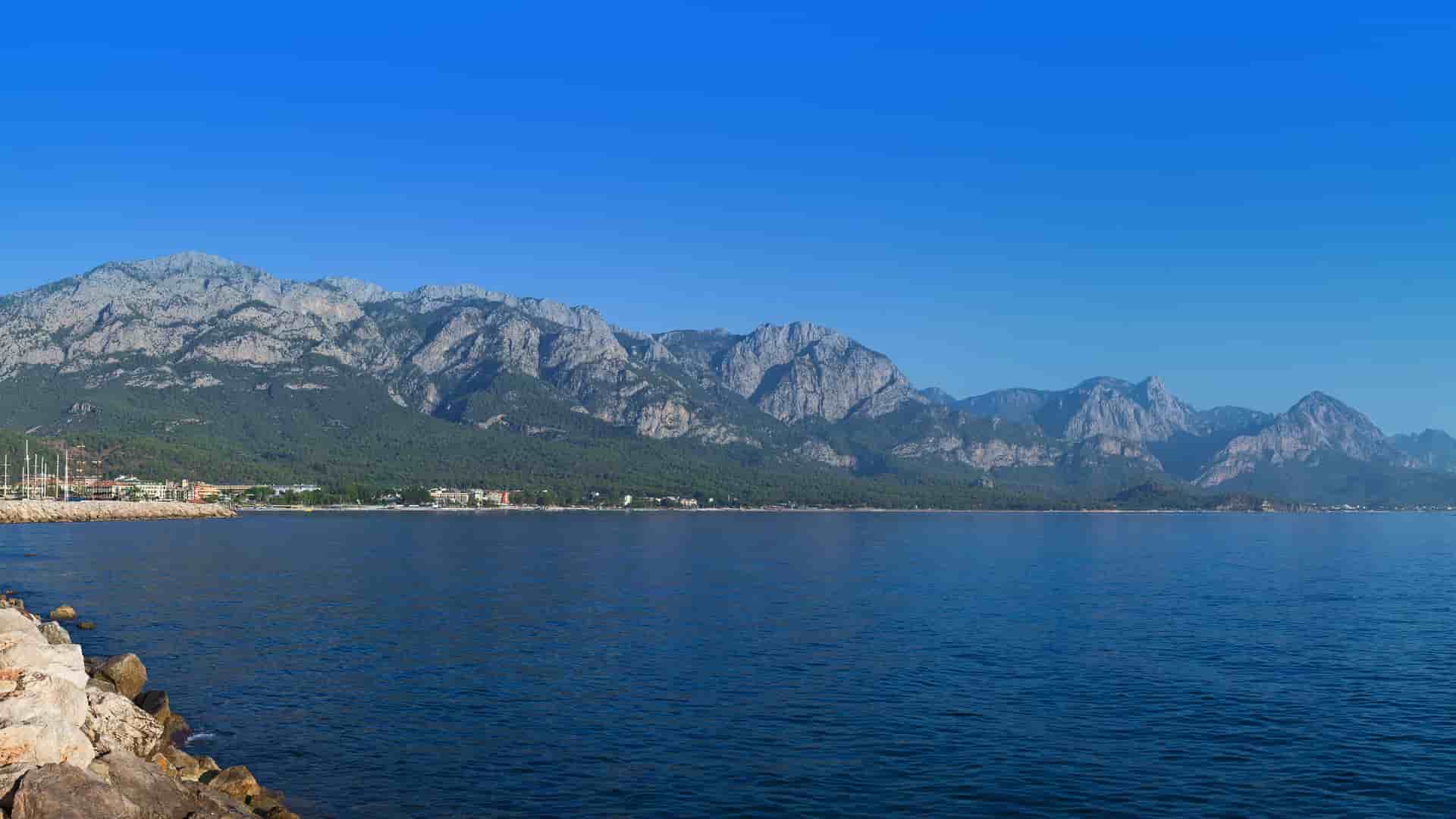 A wide panoramic view of the scenic coastal town of Dikili, Turkey, nestled on the Aegean Sea with a dramatic, rugged mountain range rising in the background.