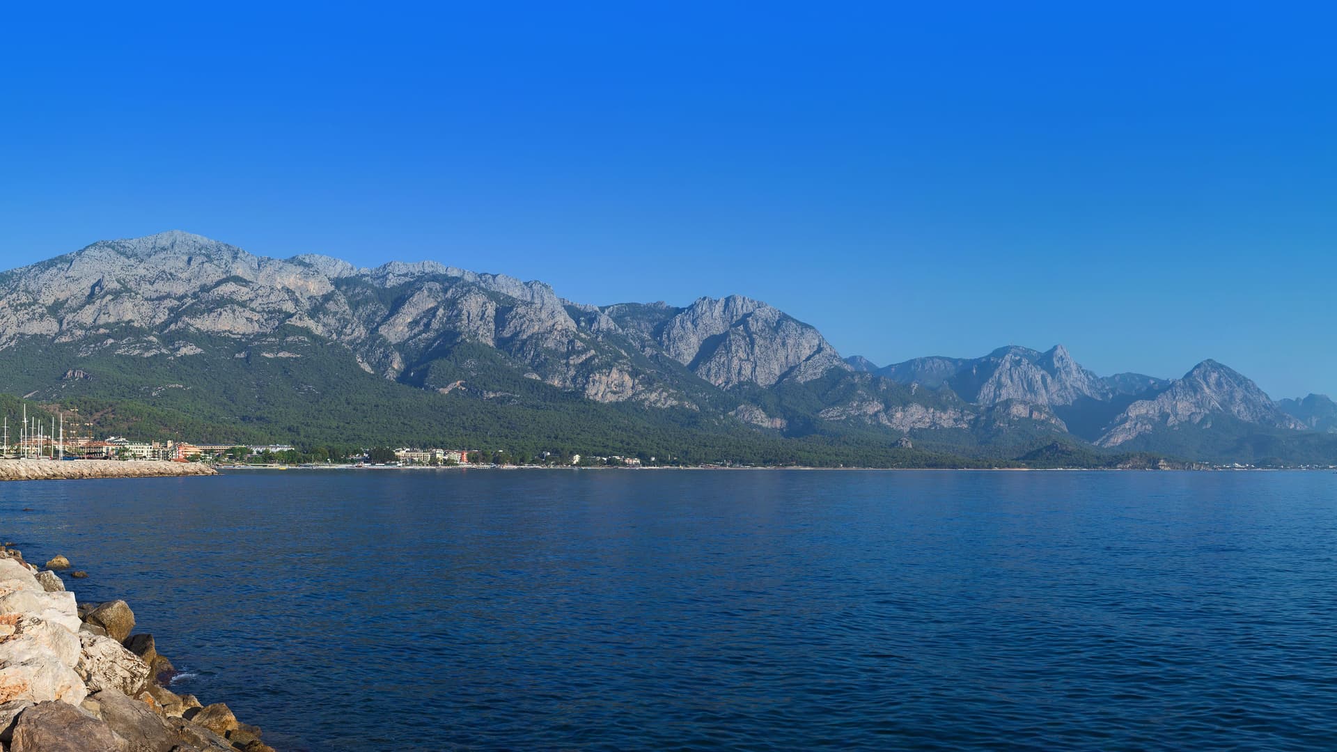 A wide panoramic view of the scenic coastal town of Dikili, Turkey, nestled on the Aegean Sea with a dramatic, rugged mountain range rising in the background.