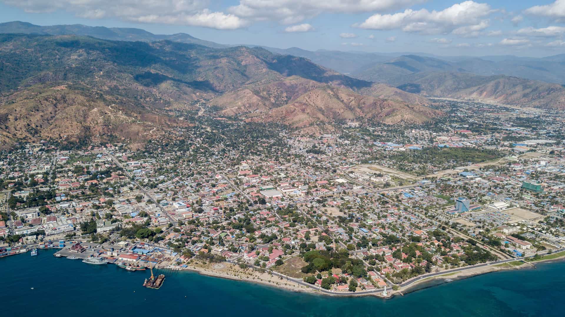 An aerial photograph of Dili, the capital city of East Timor, showcasing its bustling port, coastal urban development, and the rugged, mountainous landscape behind the city.