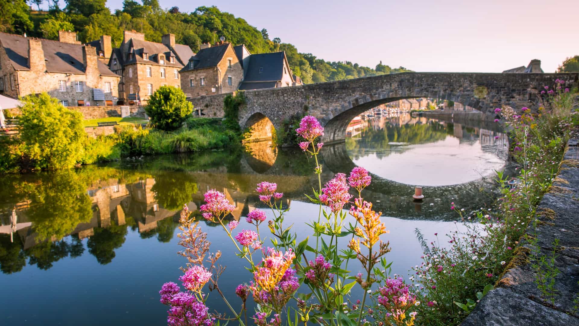 A picturesque view of the Rance River in Dinan, Brittany, France, with a historic stone bridge and medieval half-timbered houses reflected in the calm water.
