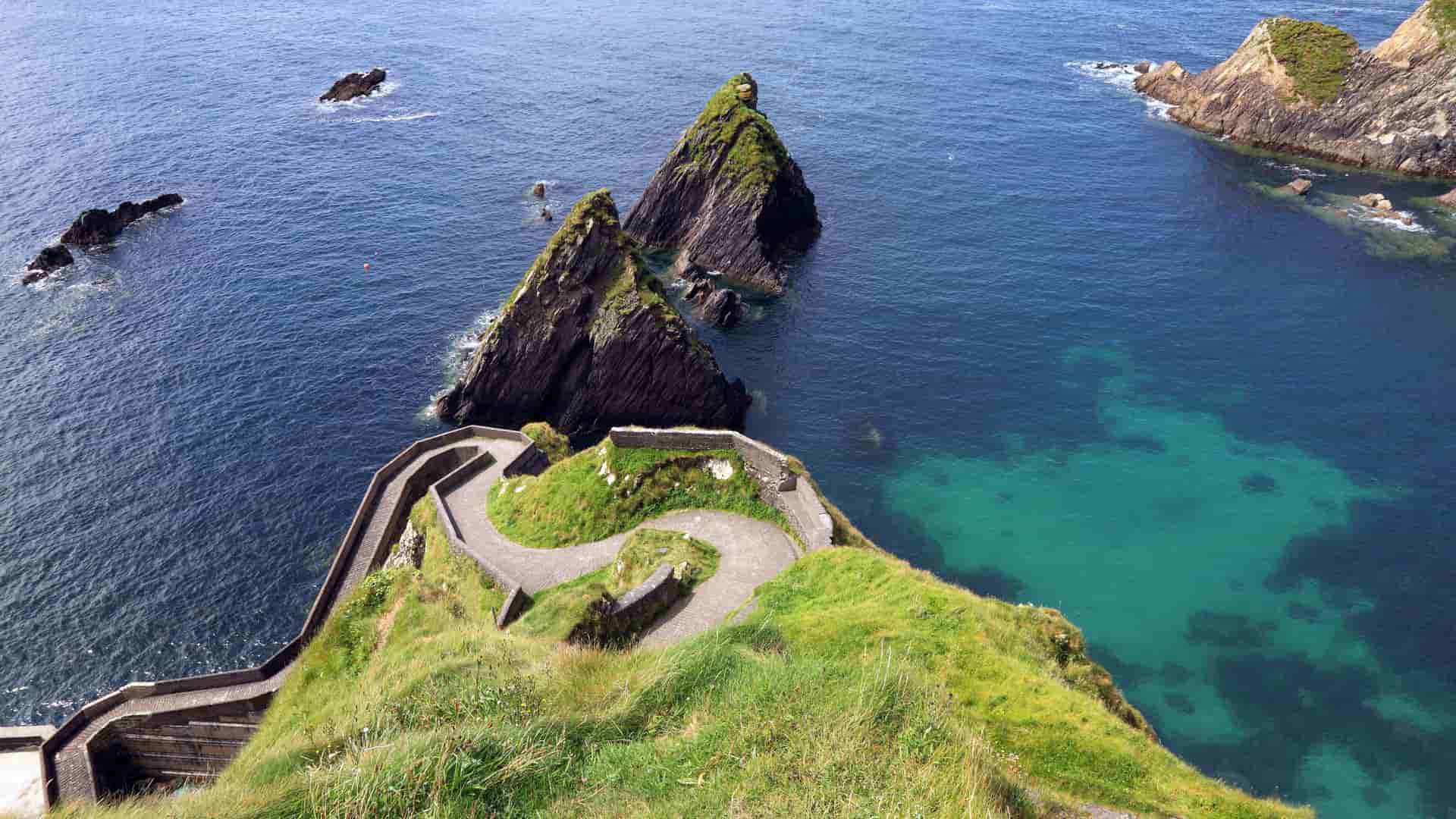 The iconic winding path and staircase at Dunquin Pier on the Dingle Peninsula, Ireland, with dramatic sea stacks rising from the clear turquoise water below.