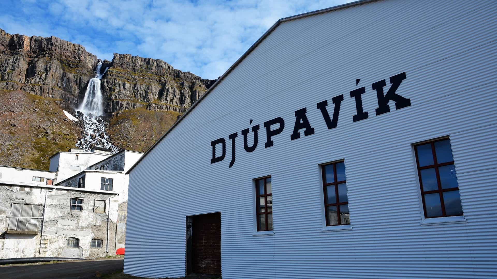 The historic abandoned herring factory in the remote village of Djúpavík, Iceland, with a powerful waterfall cascading down the dramatic, rocky cliffs behind it.