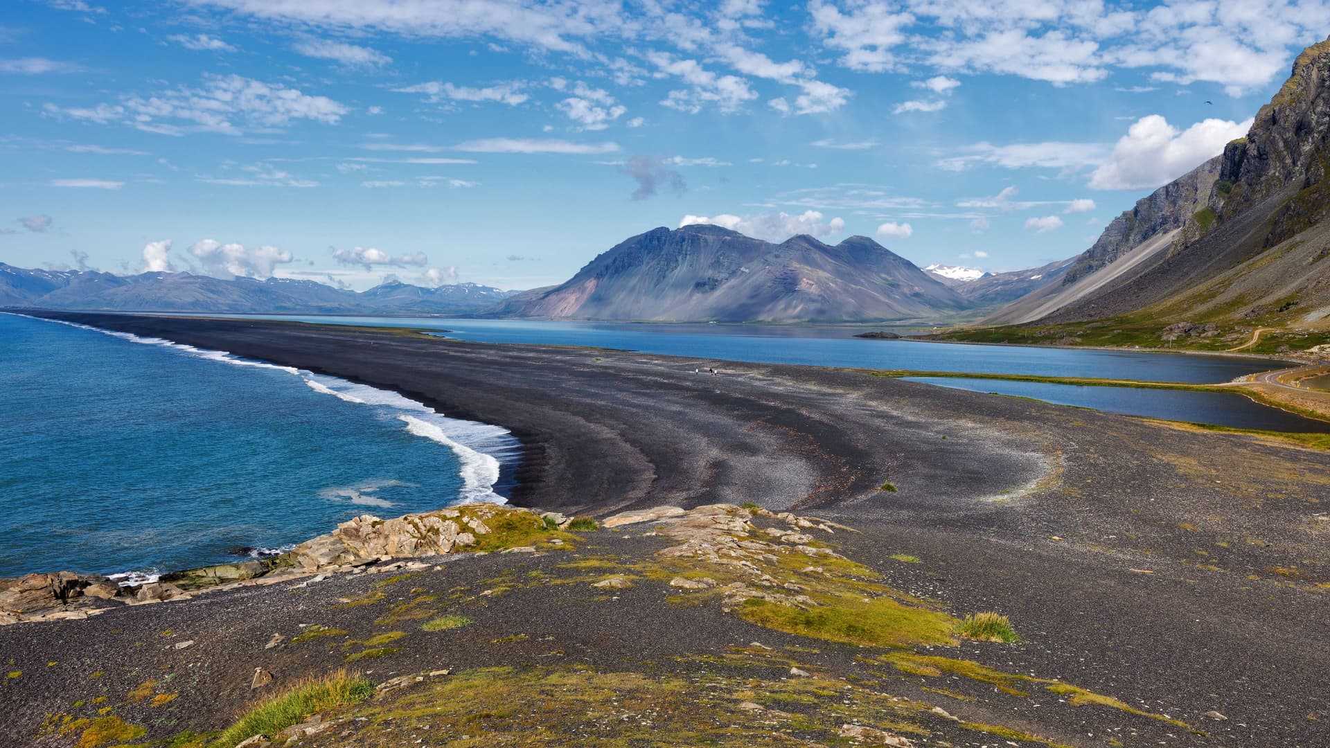 A panoramic view of the dramatic black sand beach and rugged coastline near Djúpivogur, Iceland, with a sweeping mountain range in the background.