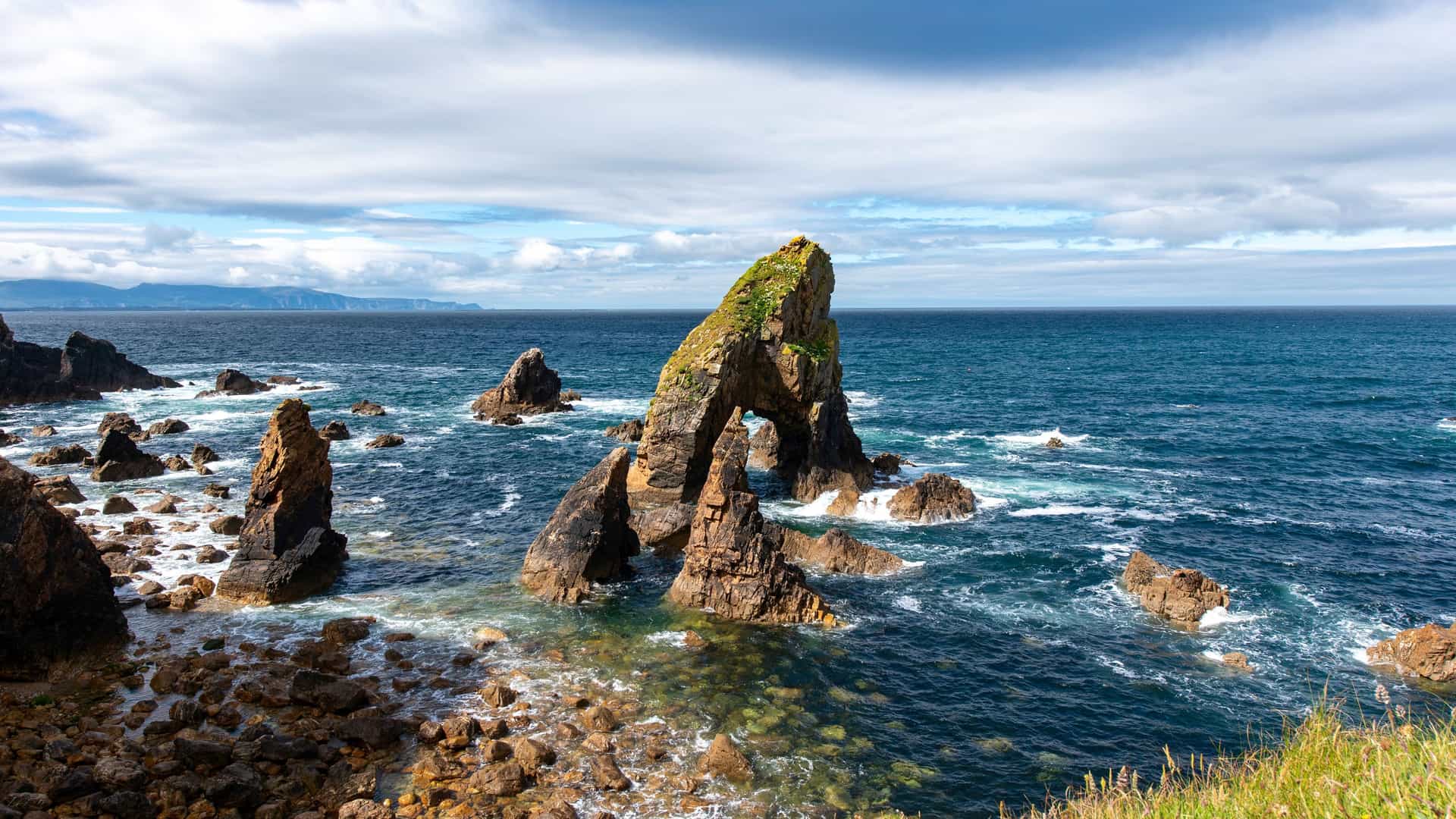 The dramatic coastline of County Donegal, Ireland, showcasing a striking rock formation with a natural sea arch and numerous sea stacks in the deep blue Atlantic Ocean.