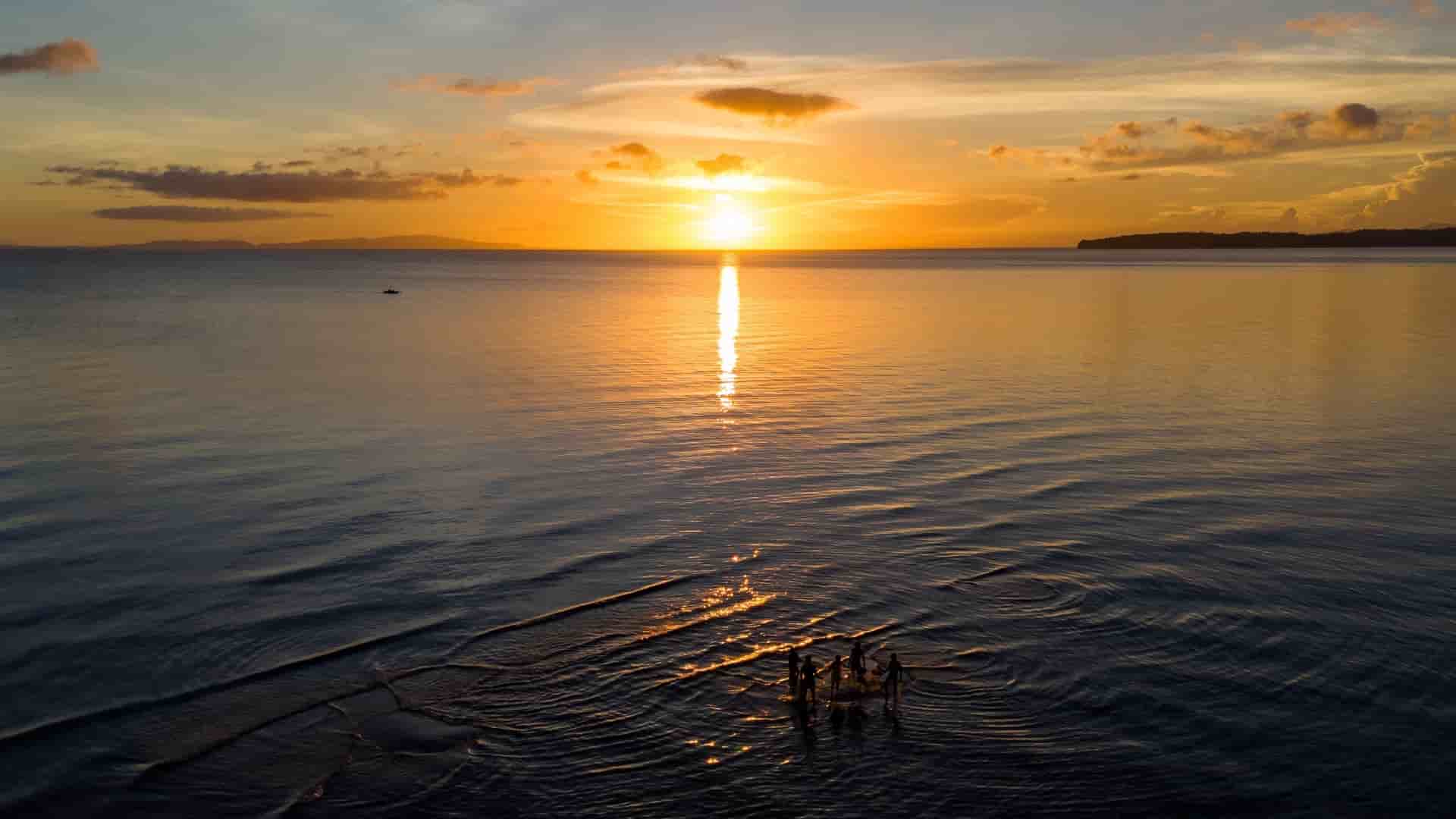 A group of people stand in the shallow water at sunset in Donsol, The Philippines, with the sun reflecting brightly.