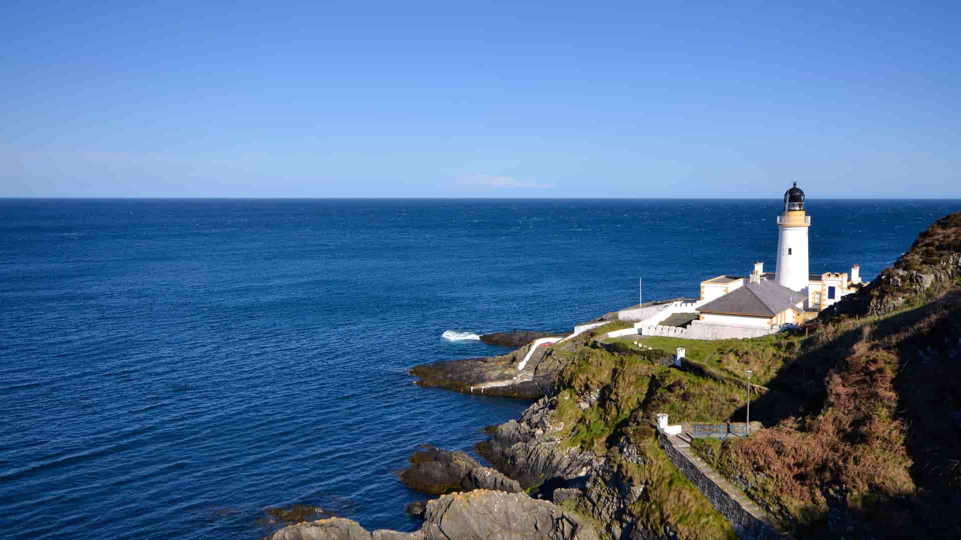 A panoramic view of Douglas Head Lighthouse on the Isle of Man, a white lighthouse situated on a green, rocky cliff overlooking the calm blue Irish Sea.