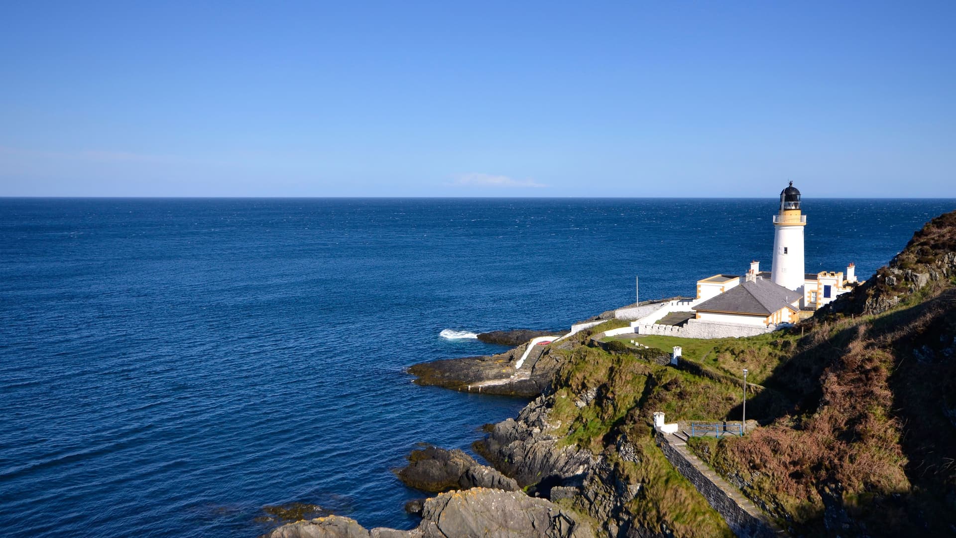 A panoramic view of Douglas Head Lighthouse on the Isle of Man, a white lighthouse situated on a green, rocky cliff overlooking the calm blue Irish Sea.