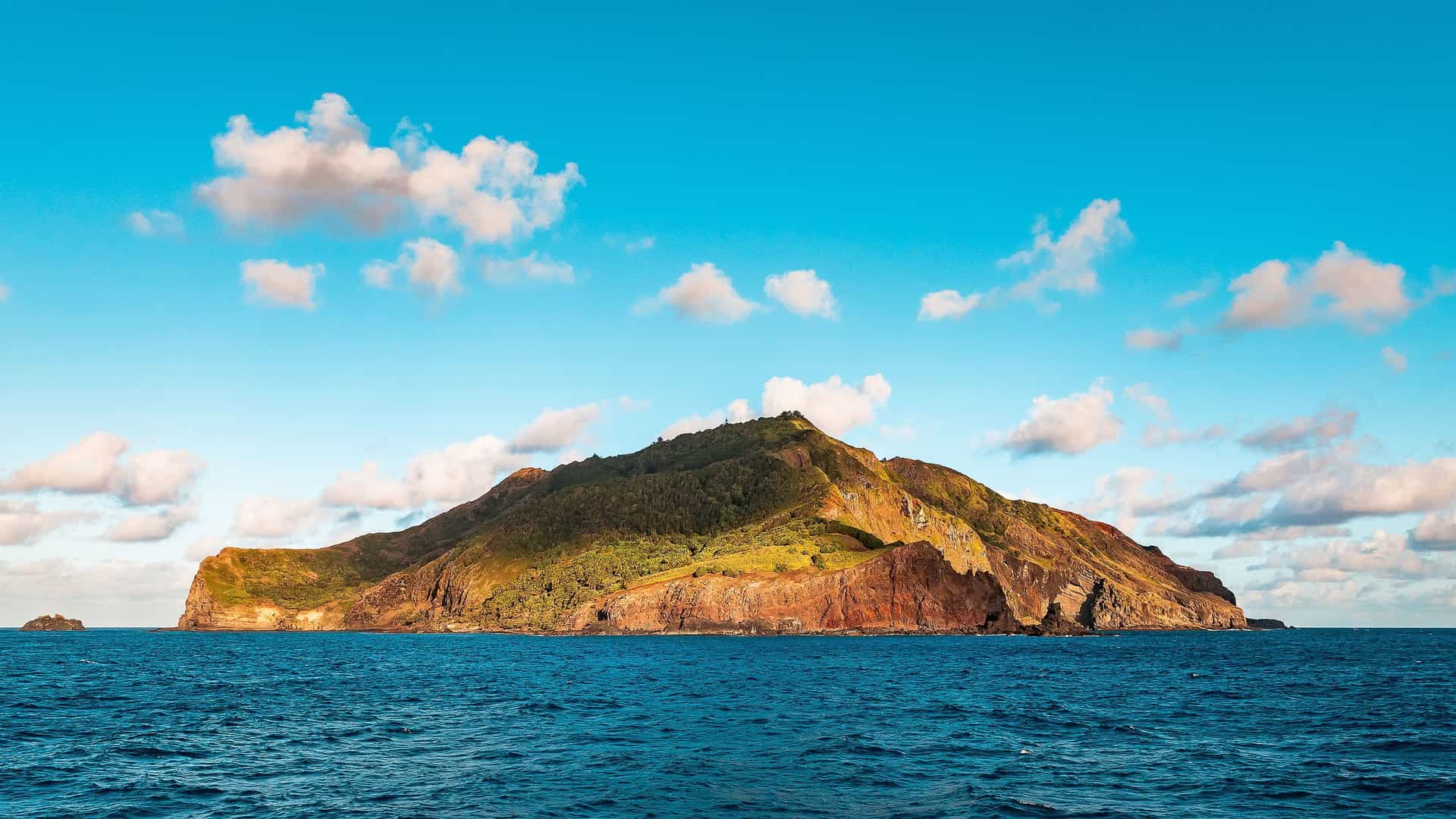 A remote and uninhabited Ducie Island with a rugged, green-covered volcanic peak rising from the deep blue ocean, under a beautiful blue sky with fluffy white clouds.