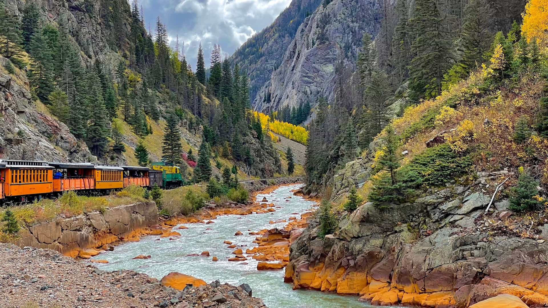 "A vibrant scene of the historic Durango & Silverton Narrow Gauge Railroad train traveling along a track beside the Animas River in a narrow canyon. The surrounding slopes are covered in pine and golden aspen trees in the autumn. "
