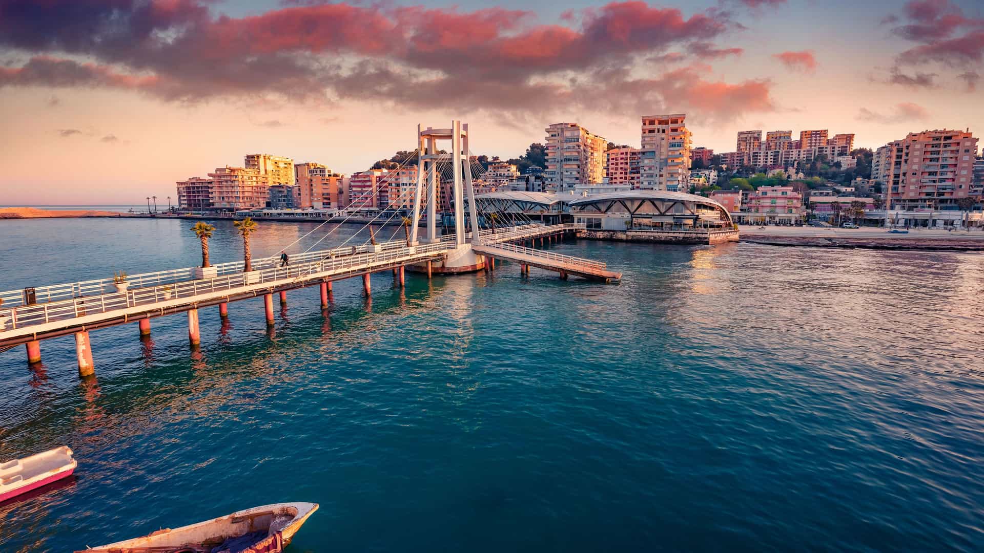 "A vibrant scenic view of the coastal city of Durrës, Albania, with a modern, white pedestrian bridge crossing the harbor toward the city's waterfront buildings at sunset. "