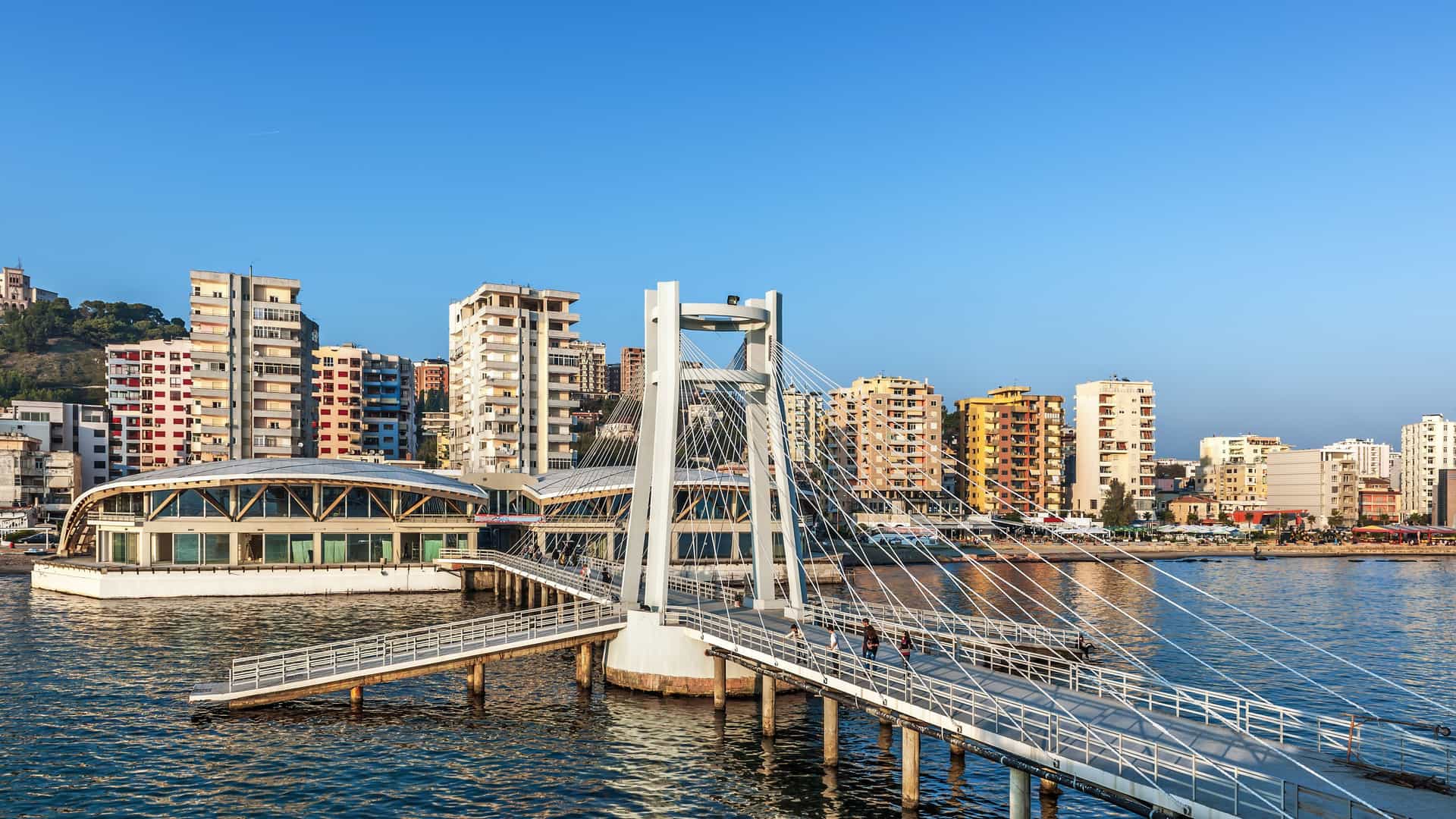 A wide-angle shot of the city of Durres, Albania, showcasing a modern pedestrian bridge stretching over the water toward colorful buildings and apartments along the harbor.