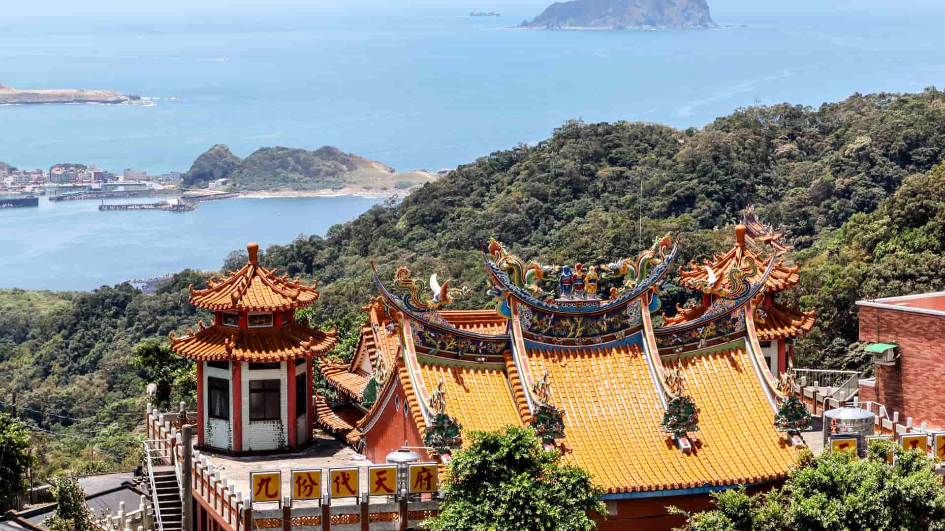 A high-angle view of a traditional Chinese temple with ornate orange and green roofs, overlooking the vast East China Sea with forested mountains and islands in the distance.