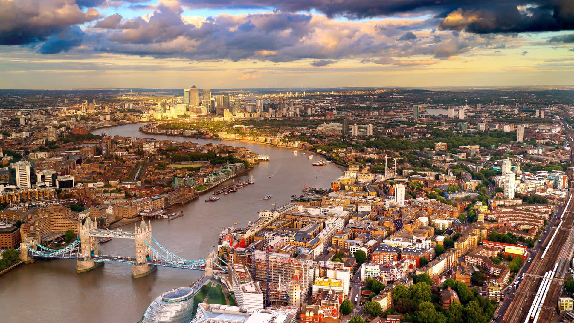 An aerial panoramic shot of East London, featuring the iconic Tower Bridge and the River Thames winding through a dense urban cityscape with modern skyscrapers in the distance.