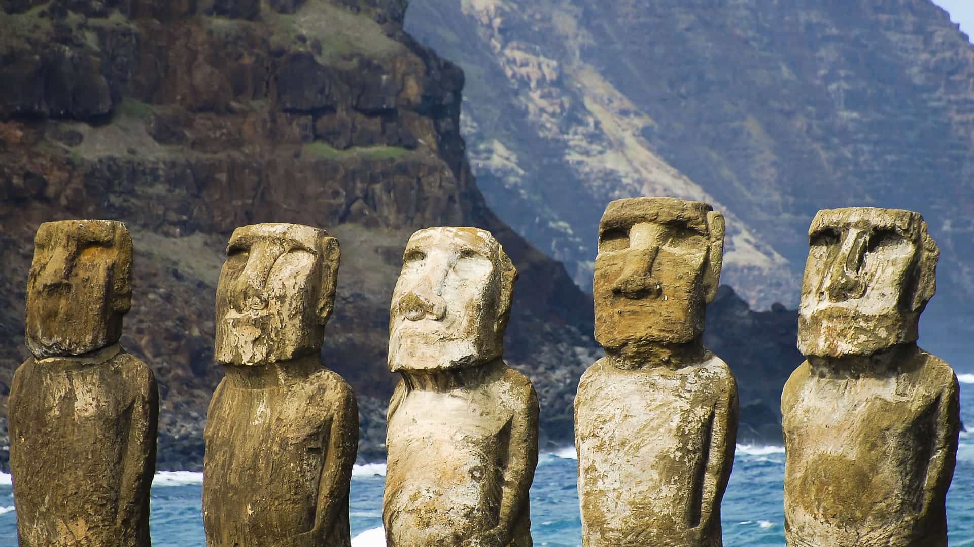 A striking lineup of several ancient Moai statues, with their unique stone faces and weathered surfaces, standing on Easter Island in front of the ocean and steep cliffs.