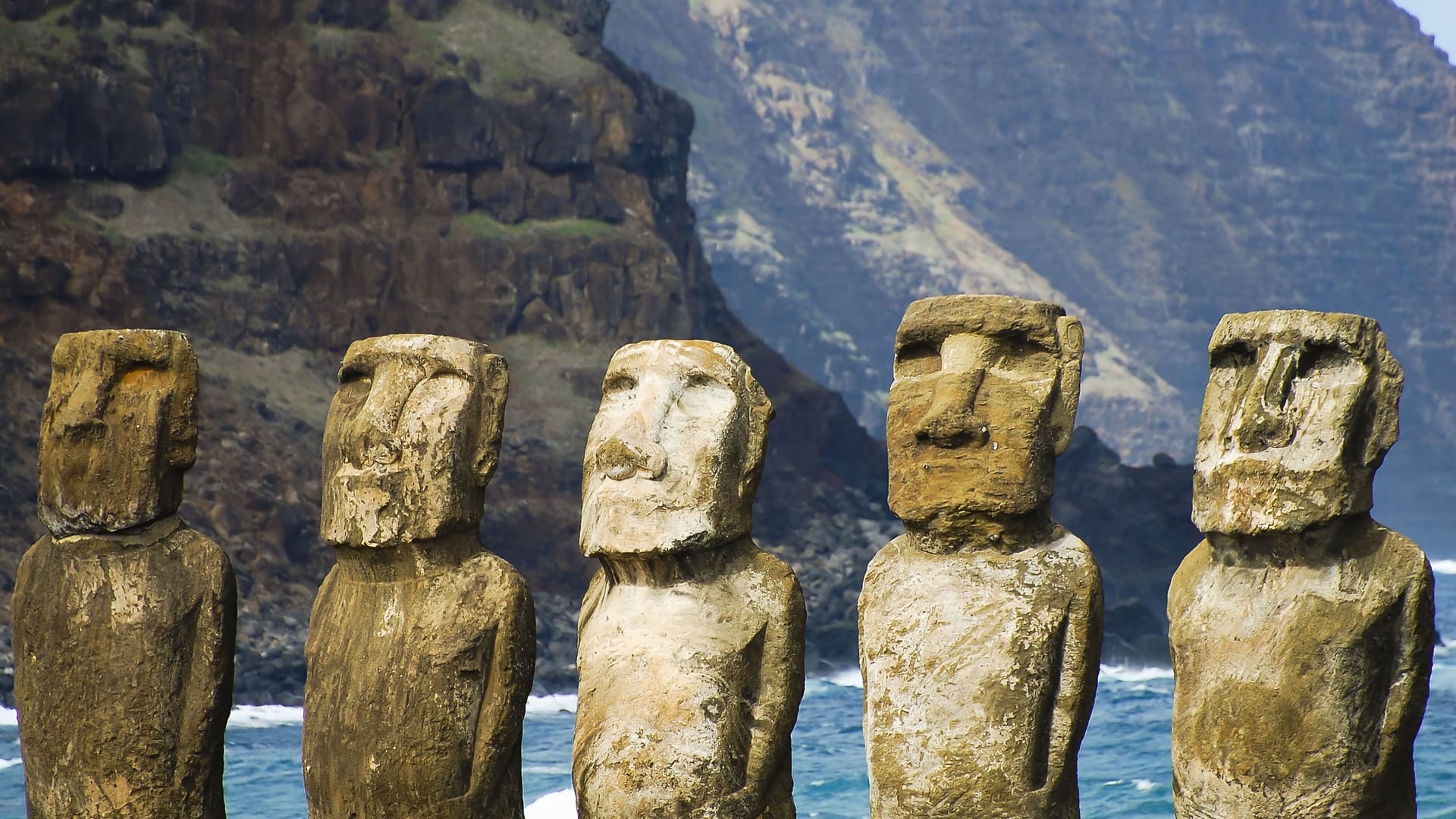 A striking lineup of several ancient Moai statues, with their unique stone faces and weathered surfaces, standing on Easter Island in front of the ocean and steep cliffs.