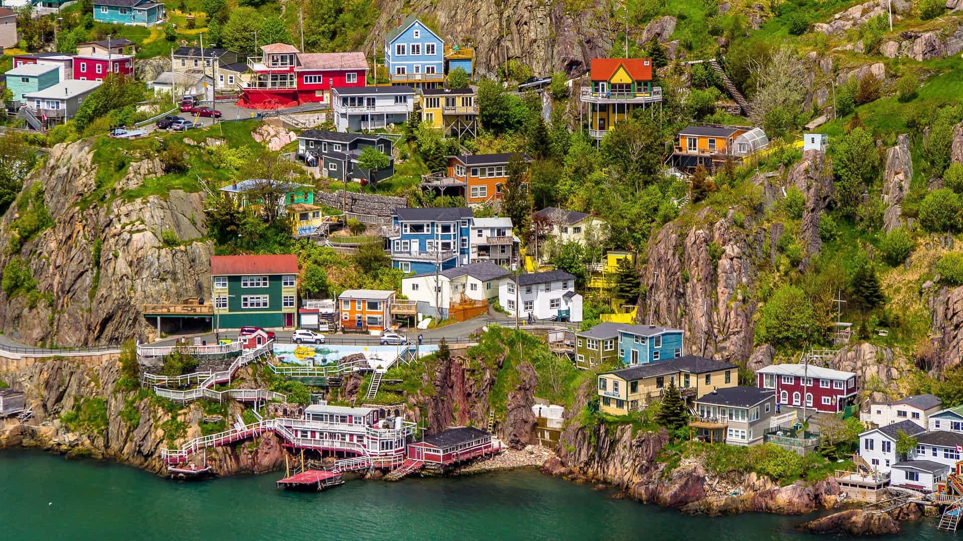 A picturesque view of St. John's, Newfoundland, showcasing a colorful collection of houses built into the rocky cliffs overlooking the deep green ocean below.