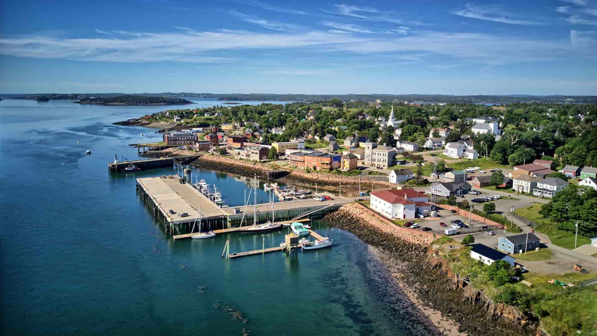 An aerial view of the coastal town of Eastport, Maine, with a busy harbor and boats docked at a long pier, surrounded by colorful buildings and lush green trees.