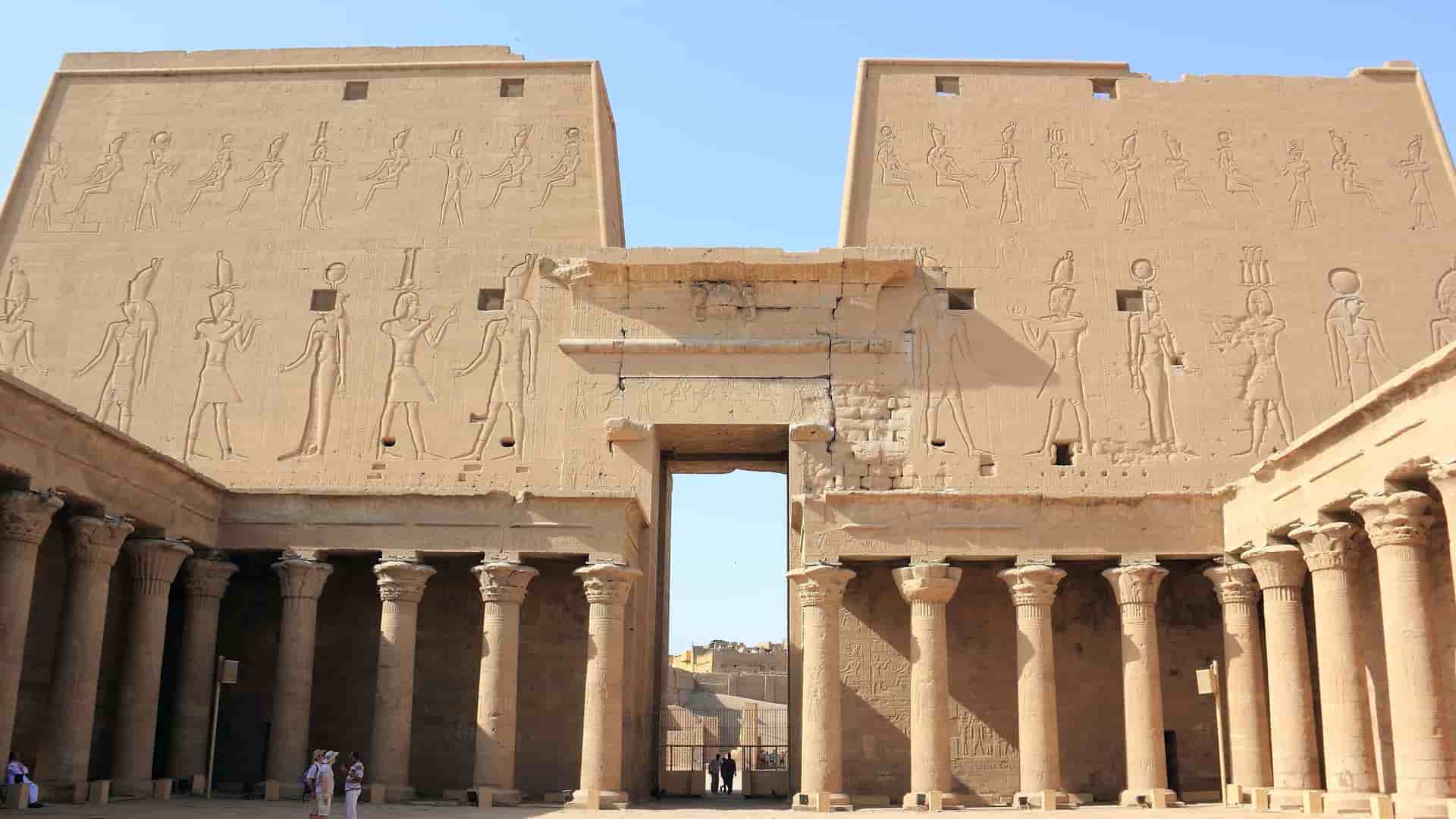 A stunning low-angle view of the ancient Temple of Horus in Edfu, Egypt, with its massive sandstone walls covered in detailed carvings and a colonnaded courtyard.