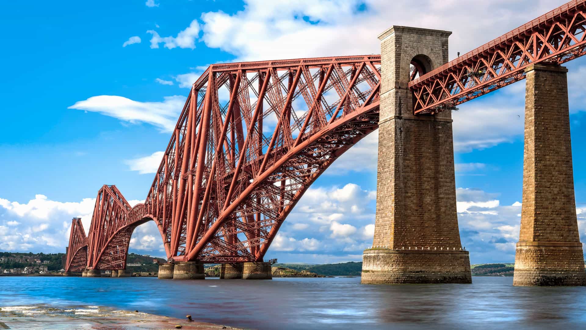 A low-angle shot of the iconic Forth Bridge near Edinburgh, Scotland, showcasing its distinctive red cantilever design and massive stone pillars stretching across the water under a bright, cloudy sky.