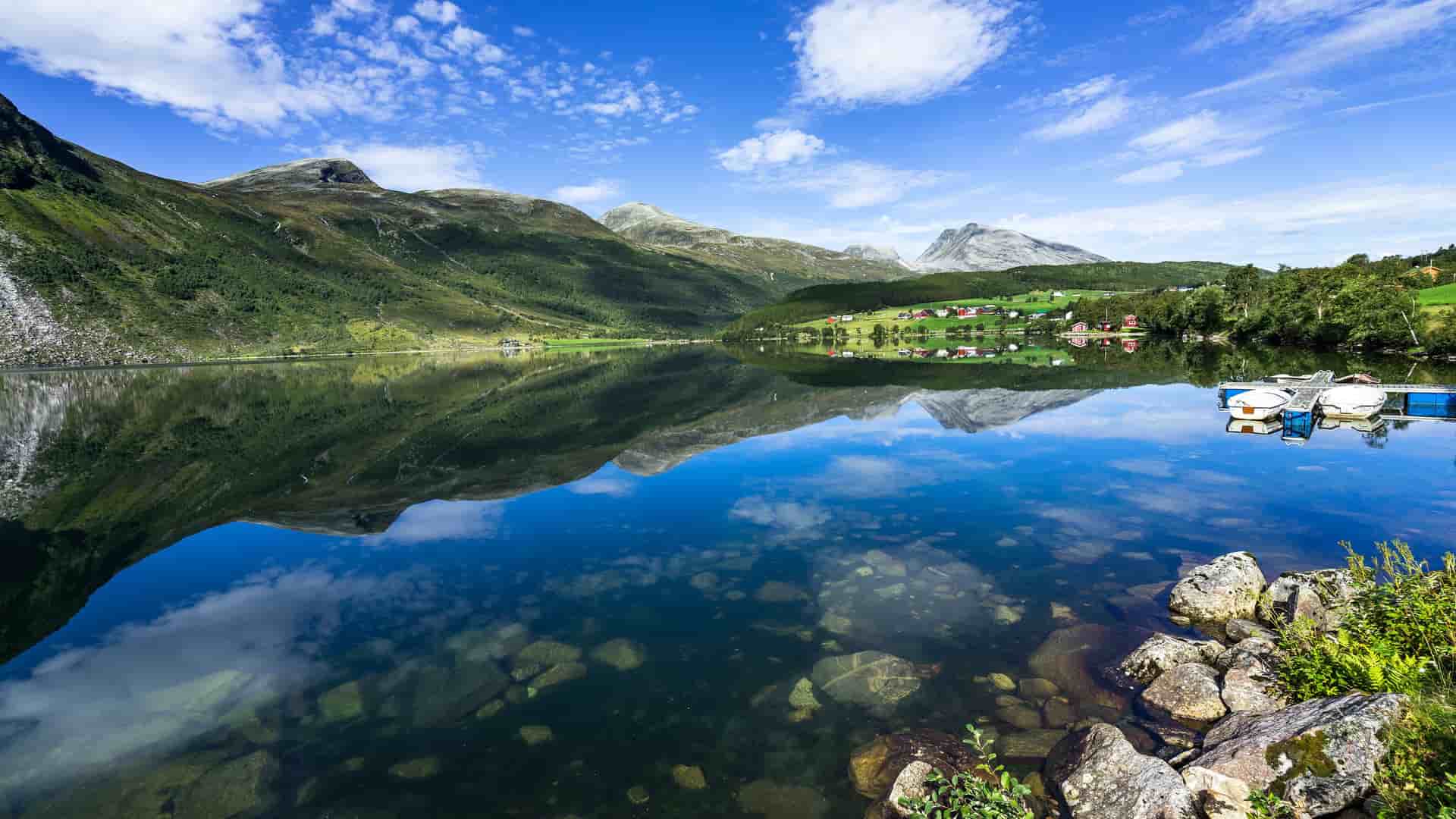"A scenic view of the tranquil lake and towering mountains of Eidsdal, Norway, with scattered houses and a small dock with boats on the far shore, all perfectly reflected in the clear, still water under a beautiful blue sky. "
