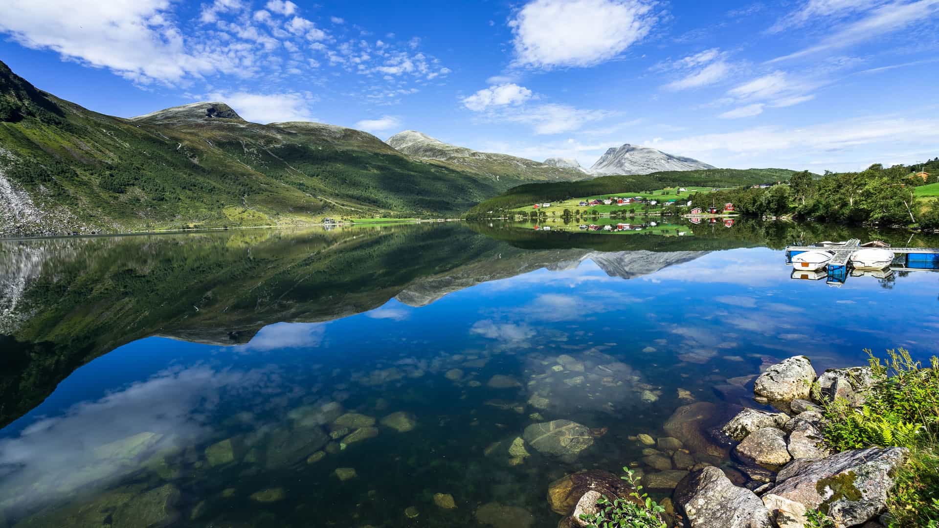 "A scenic view of the tranquil lake and towering mountains of Eidsdal, Norway, with scattered houses and a small dock with boats on the far shore, all perfectly reflected in the clear, still water under a beautiful blue sky. "