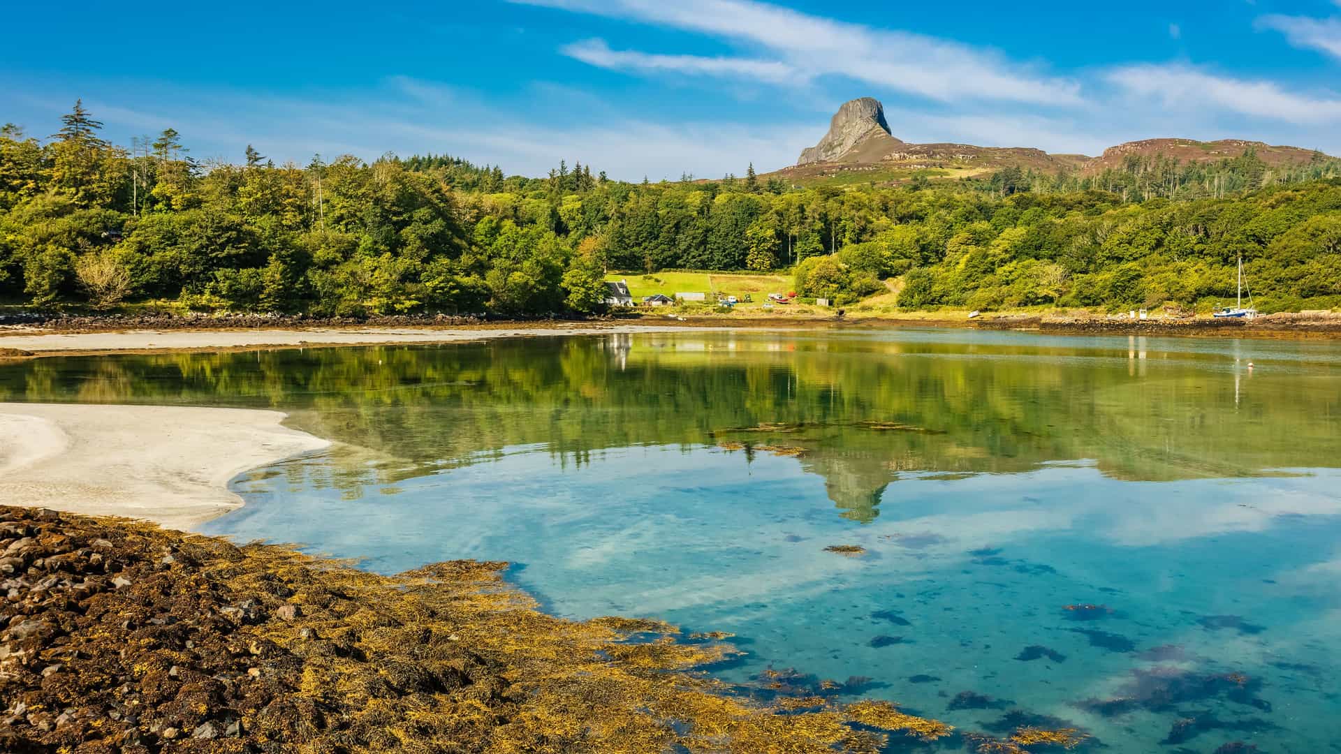 A scenic view of the Isle of Eigg, Scotland, showcasing a beautiful turquoise bay with a sandy beach and a distinctive hill, An Sgurr, in the distance, surrounded by lush green forests.