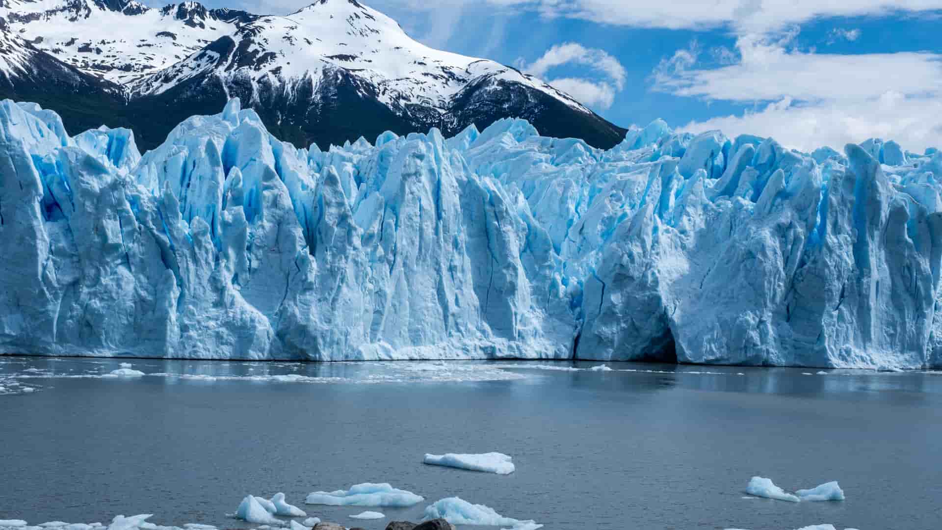 "A magnificent view of the bright blue and white face of the Perito Moreno Glacier in Patagonia, Argentina, with small icebergs floating in the frigid lake and snow-capped mountains in the background. "