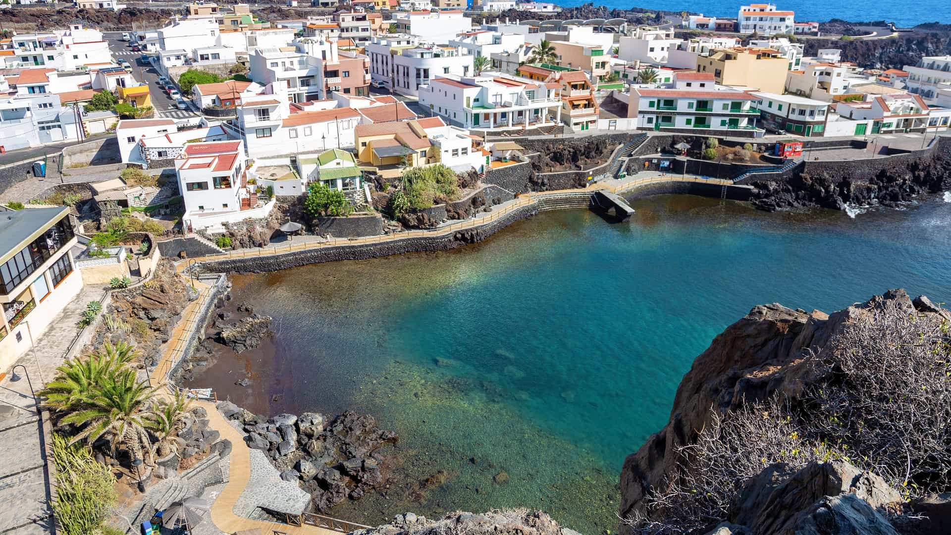 "An aerial view of the small, charming coastal town of Tamaduste on the island of El Hierro, Spain, showing its white houses and a tranquil natural swimming pool with clear turquoise water, surrounded by volcanic rocks and the blue Atlantic Ocean. "