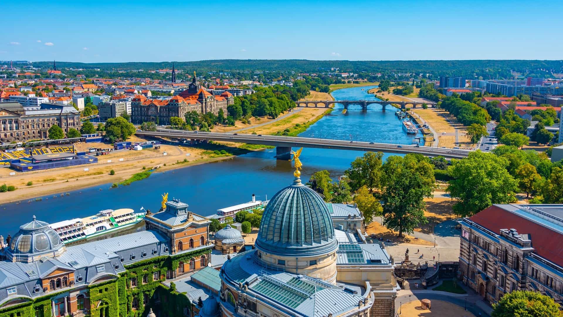 An aerial shot of Dresden, Germany, with the Elbe River flowing through the city, showcasing historic buildings, bridges, and lush green riverbanks under a clear blue sky.