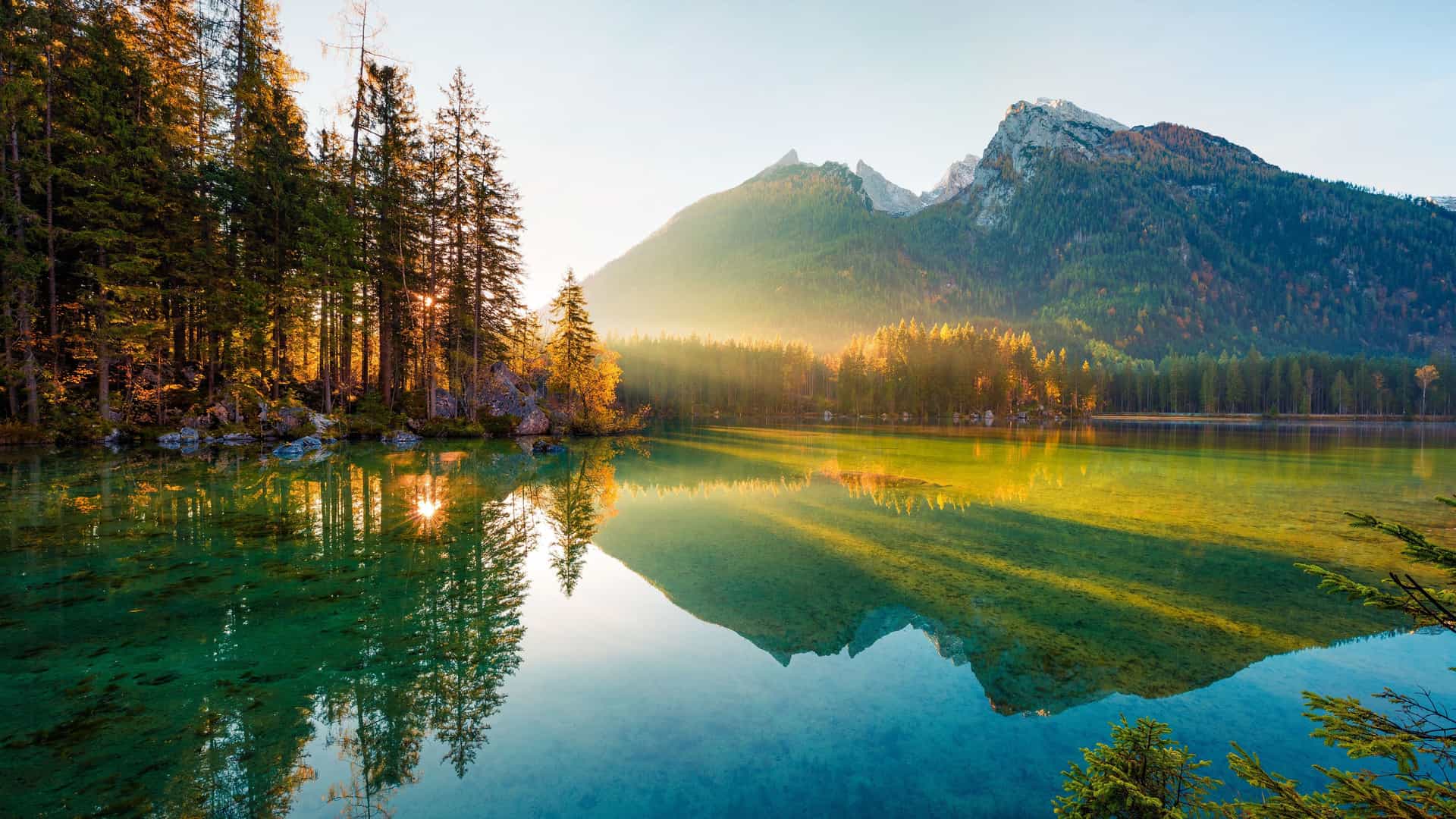 A serene sunset over a calm lake in Eltmann, Germany, with sunlight streaming through a forest of tall trees and a majestic mountain reflecting on the crystal-clear water.