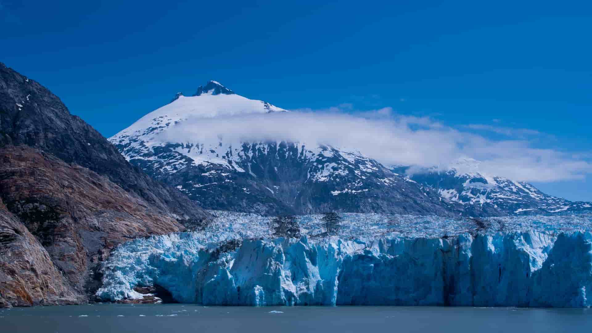 A dramatic view of a towering blue glacier and a jagged, snow-capped mountain at Endicott Arm, Alaska, with clear blue water and sky in the stunning landscape.